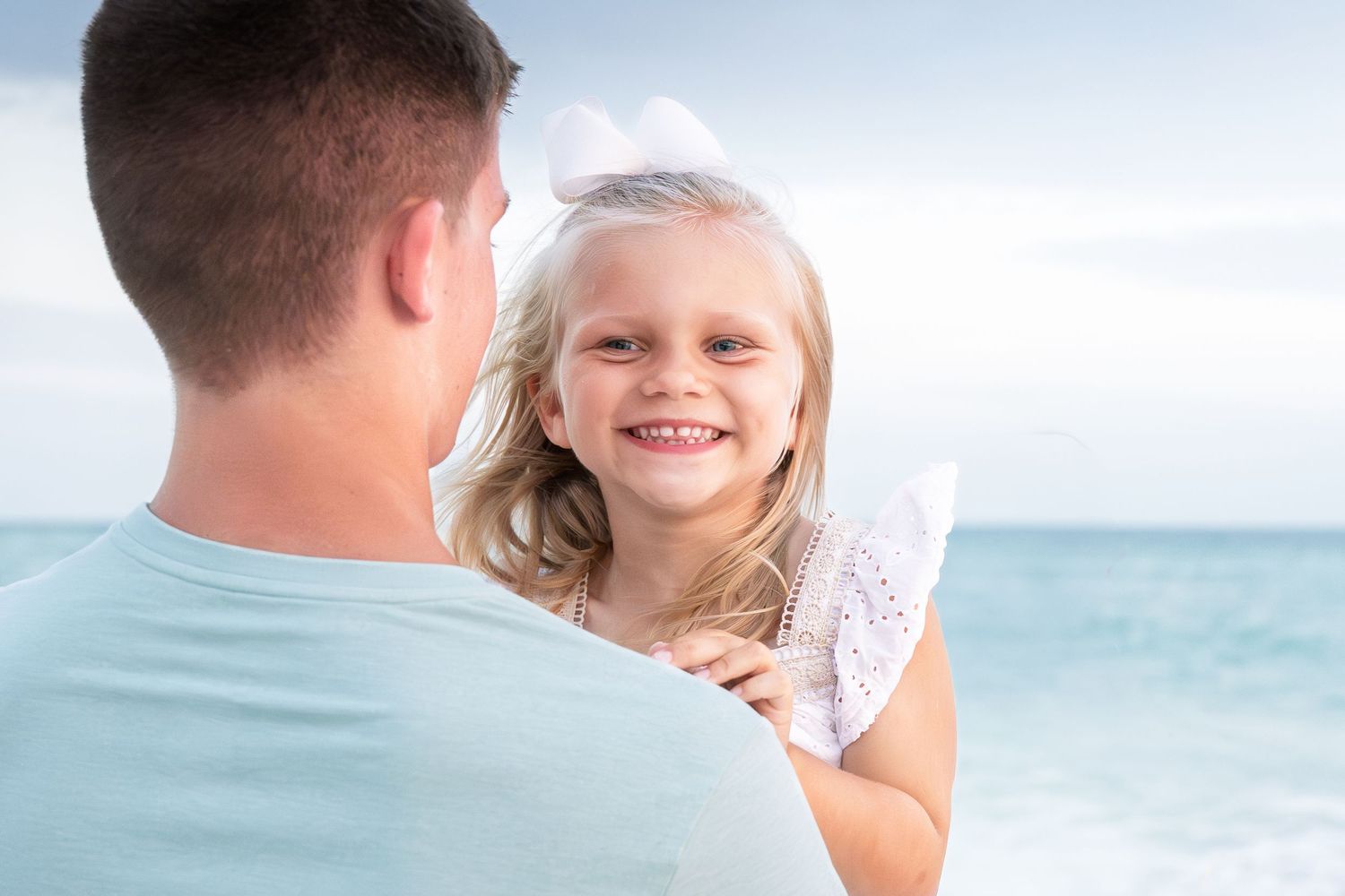 A smiling little girl with a white bow being held by her father on a sunny beach.