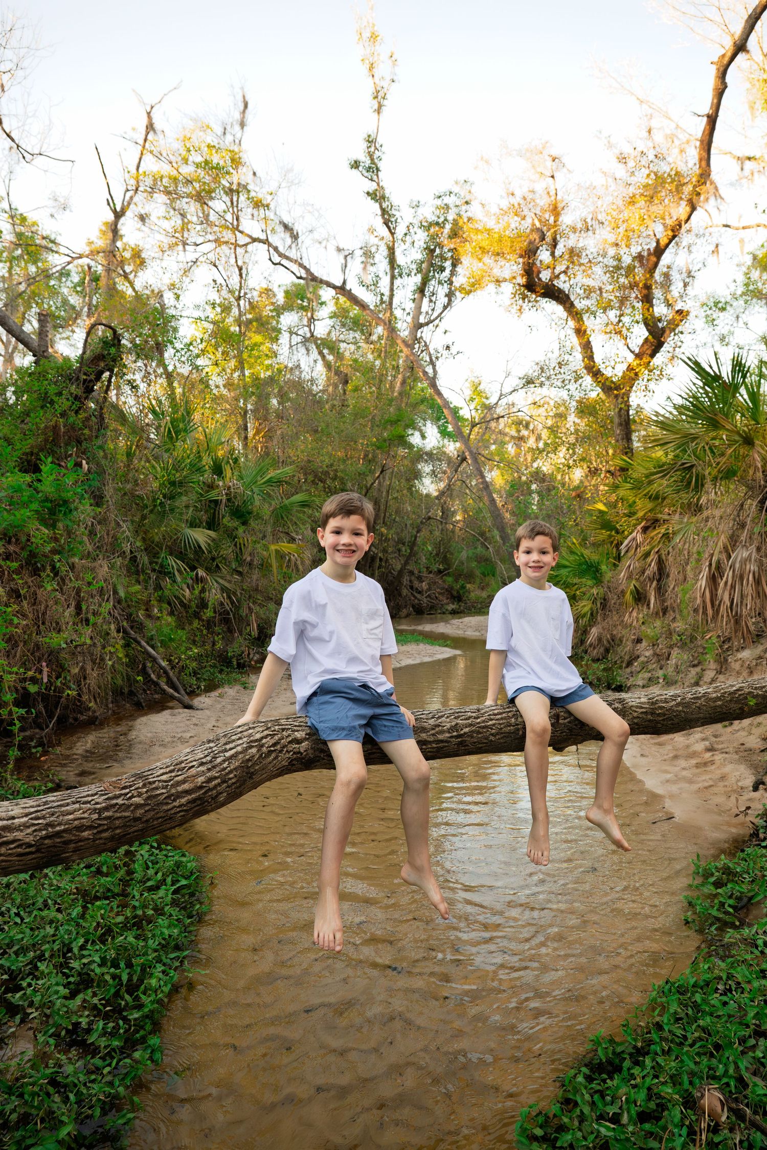 Two smiling boys in white shirts sit barefoot on a fallen log over a creek surrounded by lush green trees.