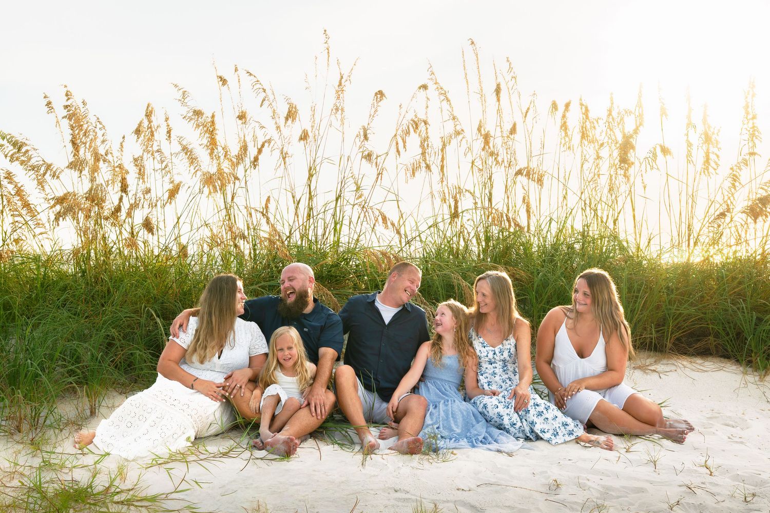Family of seven sitting together on sandy beach dunes with golden sea oats and green grass at sunset.