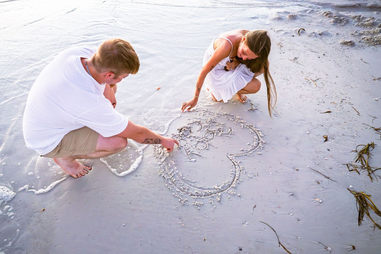 Couple drawing a heart in wet beach sand together, wearing white outfits on a romantic seaside getaway.