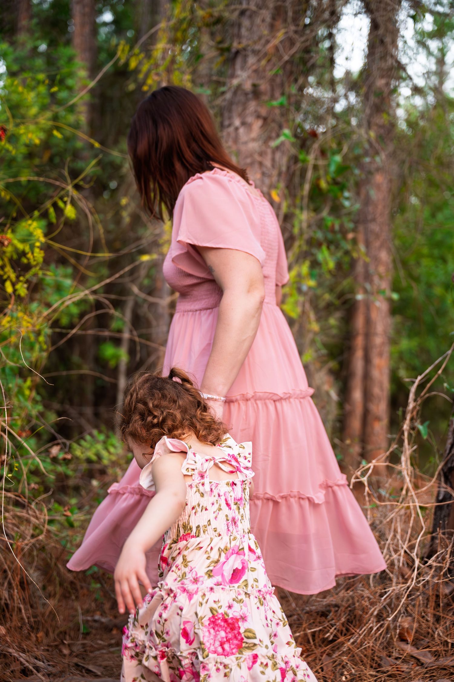 Two people in pink dresses walk through a wooded forest setting with sunlight filtering through the trees.