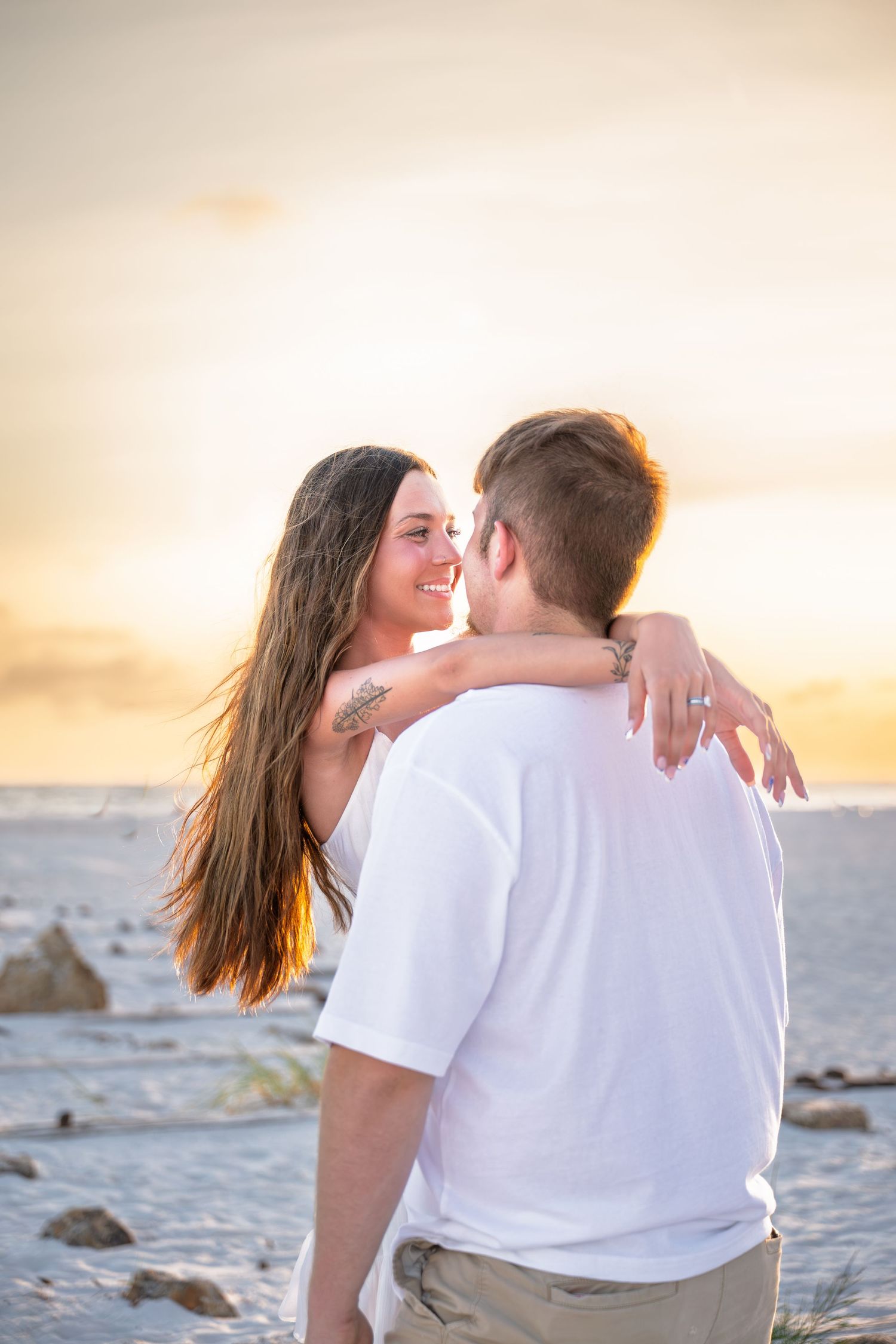 Couple embracing on beach at sunset, woman smiling with long hair and tattoo, man in white shirt, golden sky backdrop.