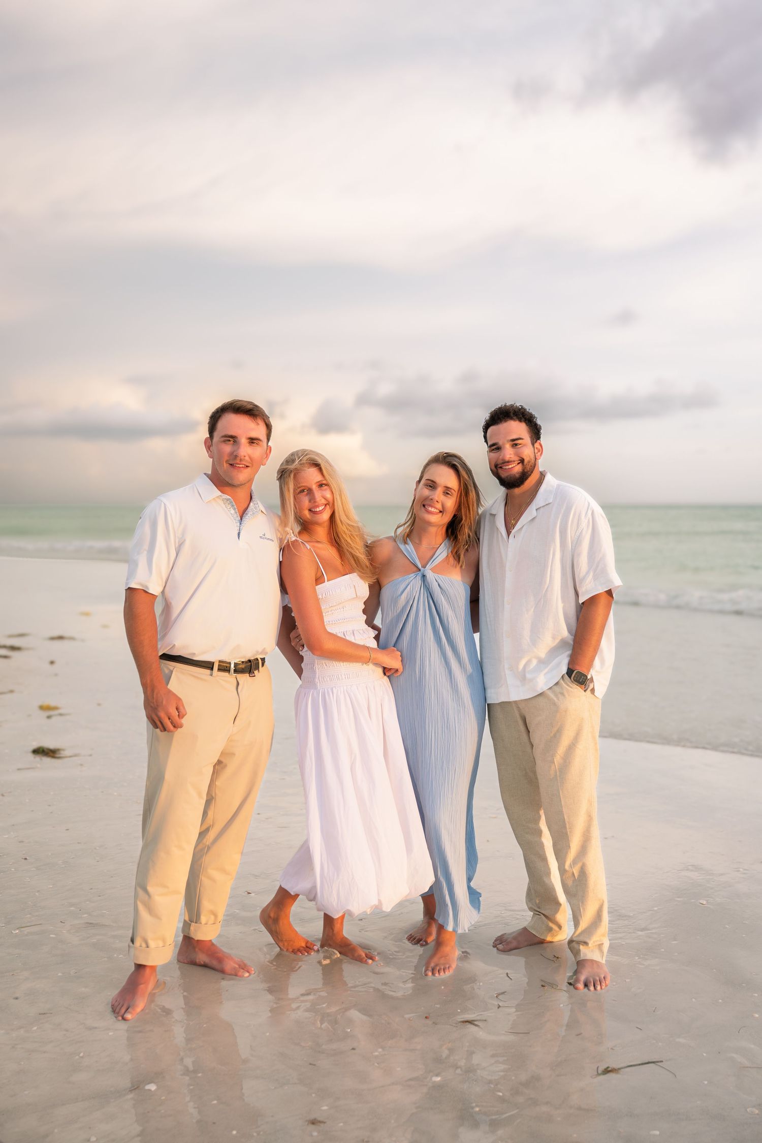 Four friends smiling together on a sandy beach at sunset, wearing casual white and blue outfits with ocean waves behind them.