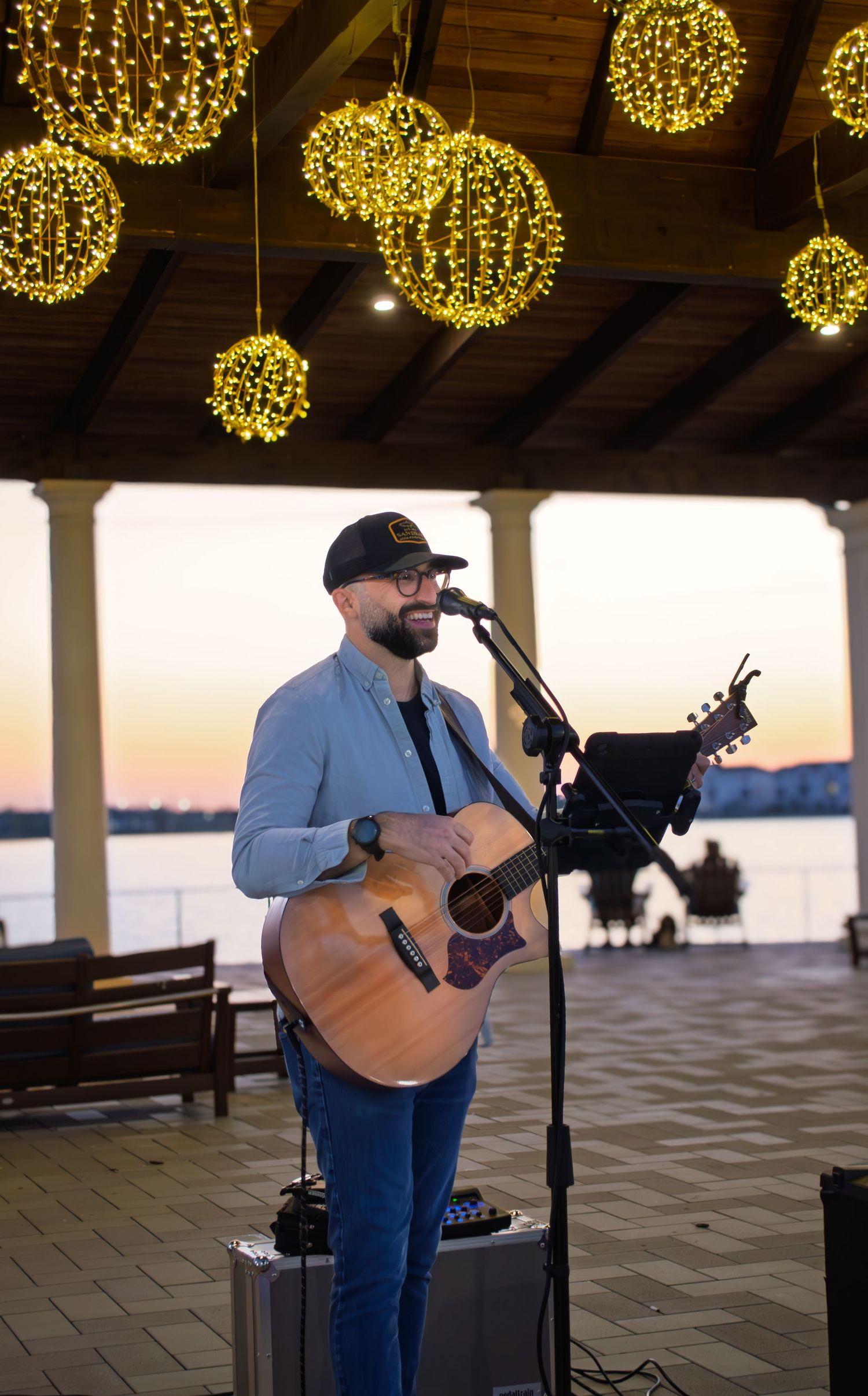 Musician performs acoustic guitar under glowing spherical lights at sunset by the waterfront.