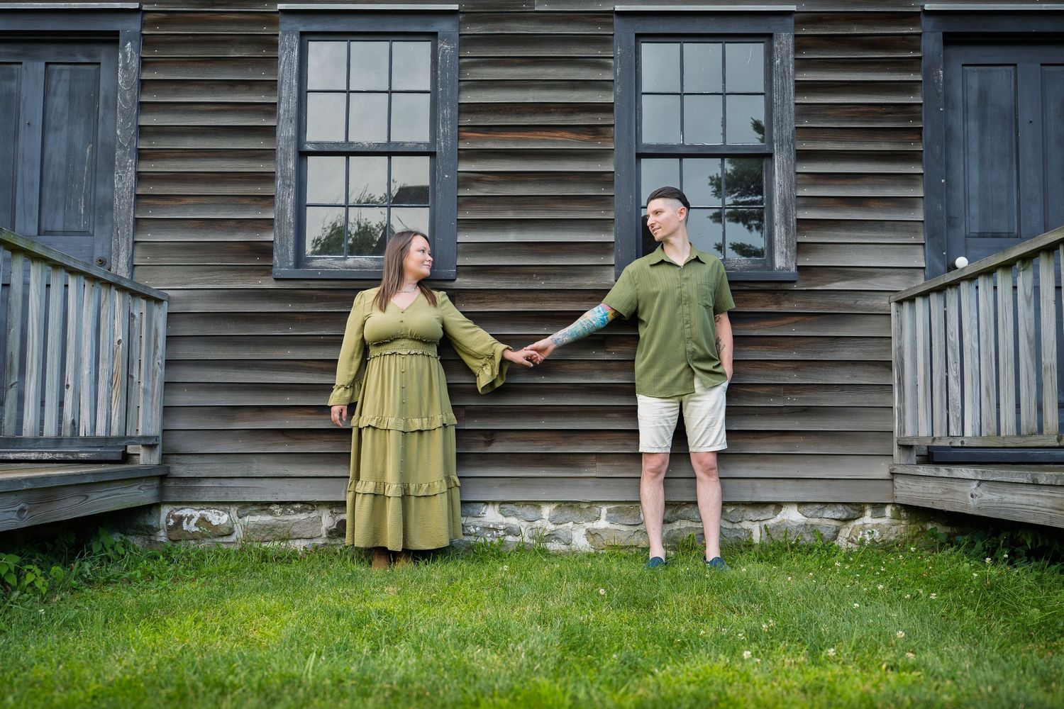 Couple in matching green outfits holding hands in front of a rustic wooden building on a sunny day.
