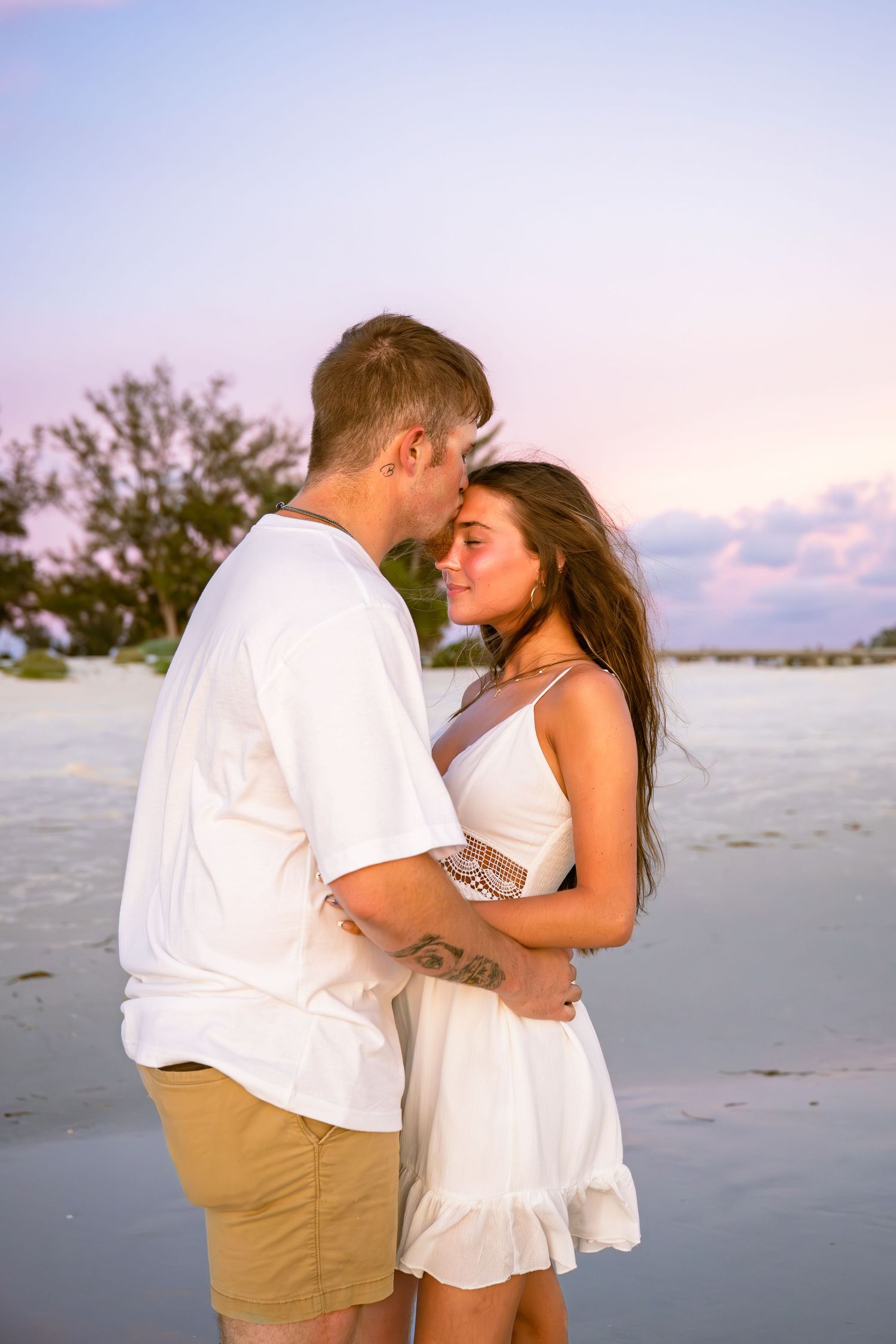 Romantic couple in white outfits embracing on a tropical beach at sunset with pink and purple sky.