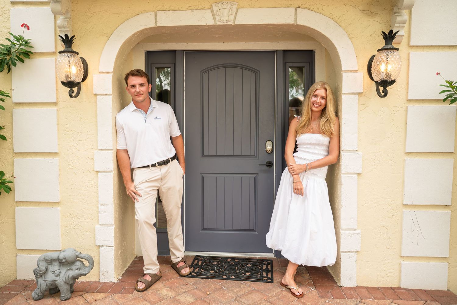 A smiling couple stands at the entrance of an elegant home with a gray front door and decorative wall sconces.