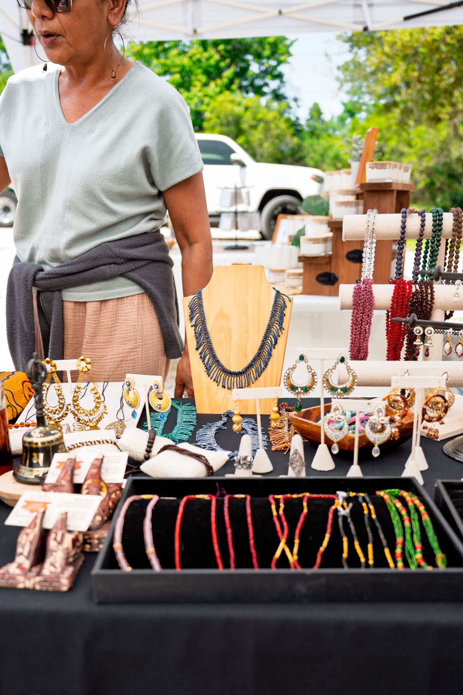 Woman vendor selling handmade jewelry at outdoor market with necklaces, earrings, and beaded accessories displayed on black table.