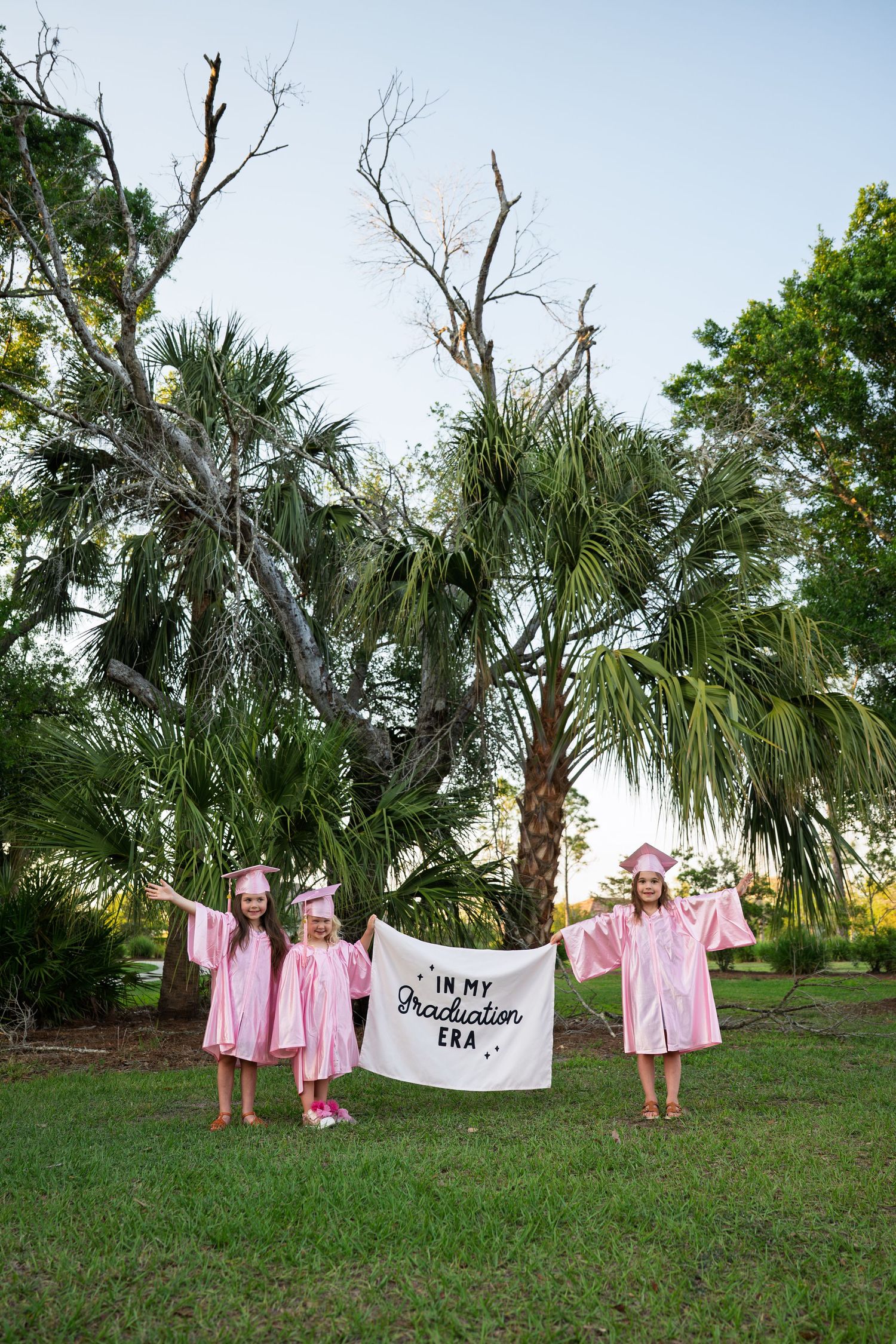 Three girls in pink graduation caps and gowns holding an 'In My Graduation Era' banner outdoors.