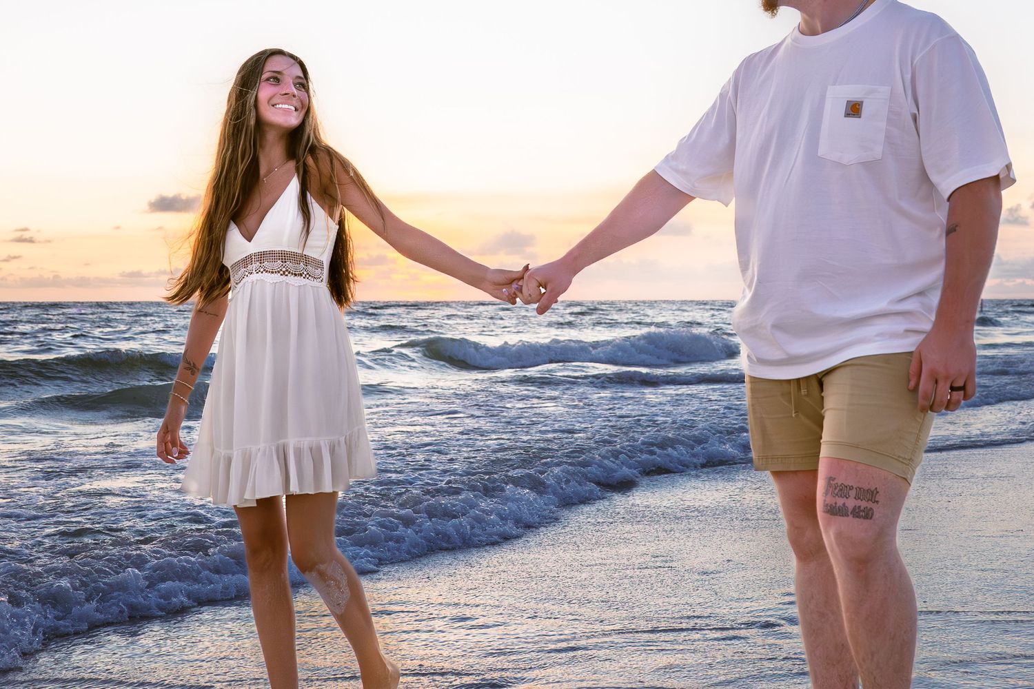 A couple holds hands walking along a sandy beach at sunset, woman in white dress, man in khaki shorts and white tee.
