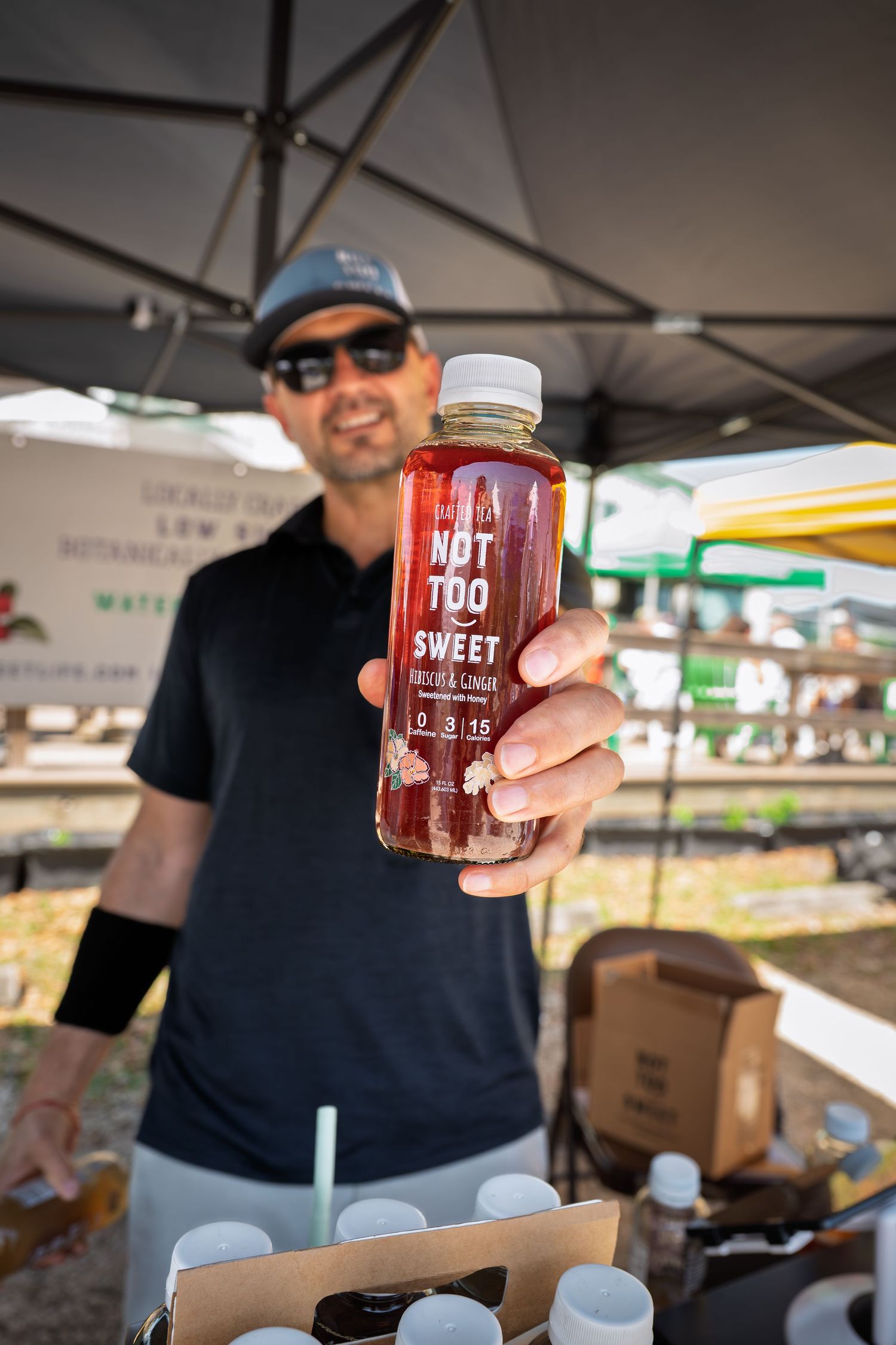 Man holding a Not Too Sweet Rose and Ginger botanical drink bottle at an outdoor farmers market vendor booth.
