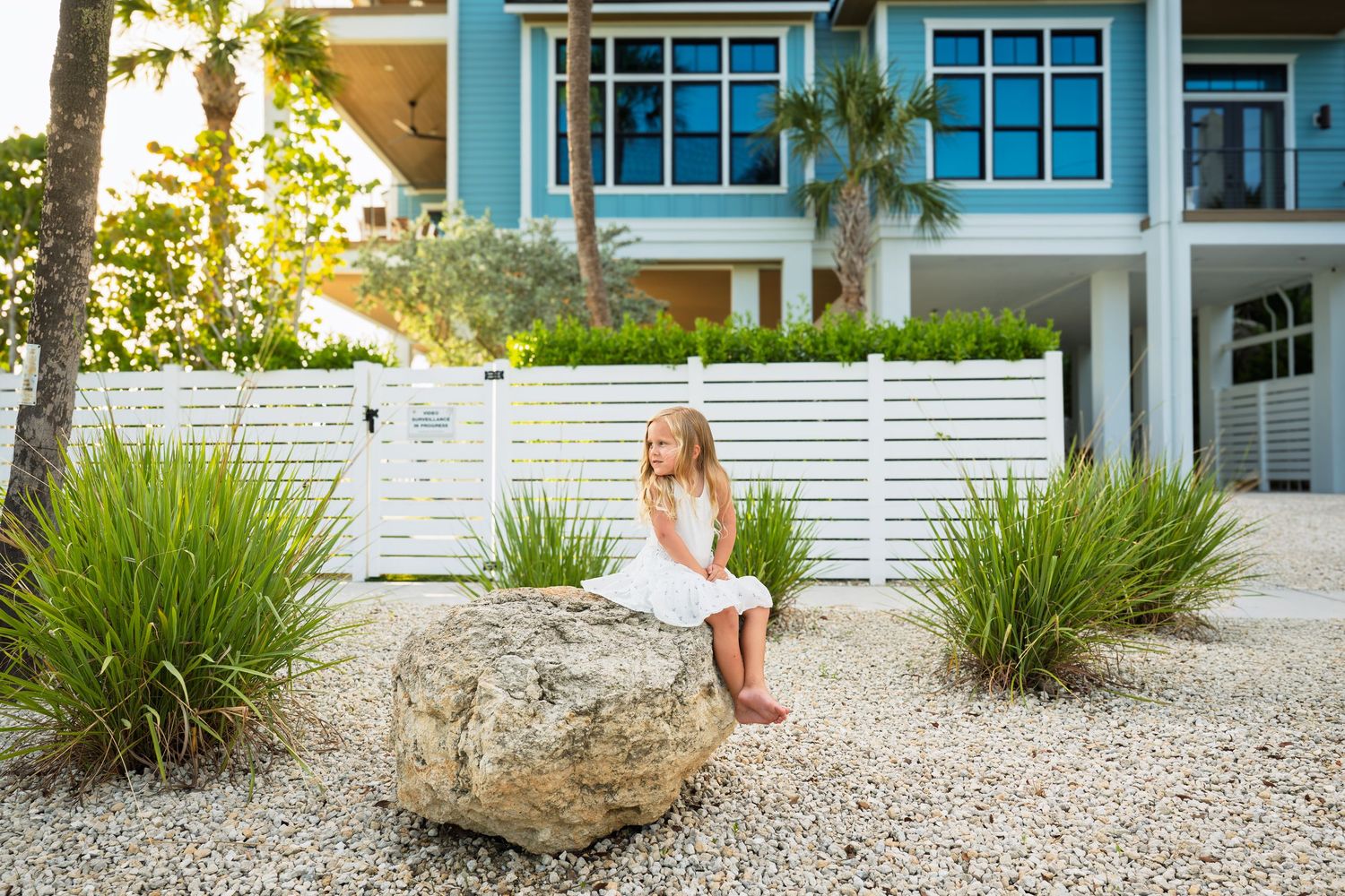 A young girl in a white dress sits on a large rock in a tropical garden outside a blue beach house.