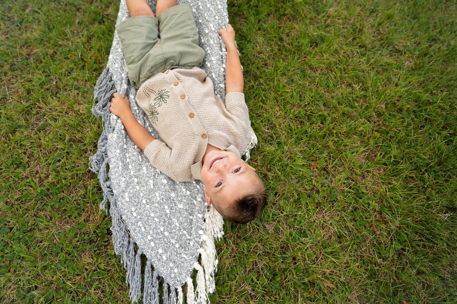 Smiling child lying on a patterned blanket with fringe on green grass, viewed from above.