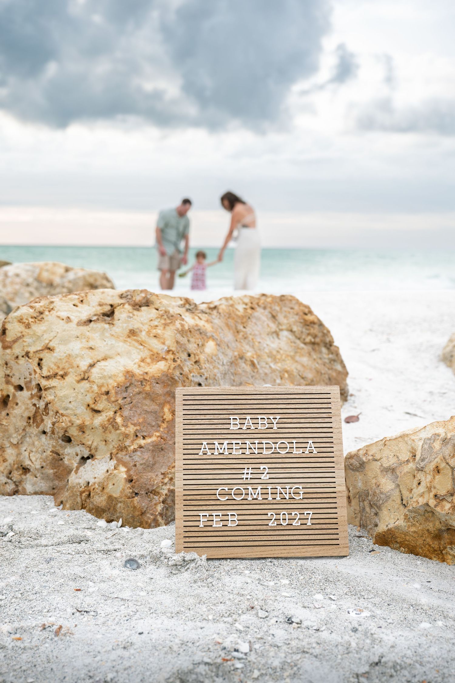 Beach pregnancy announcement sign reading Baby Amendola #2 Coming Feb 2027 with family in background.