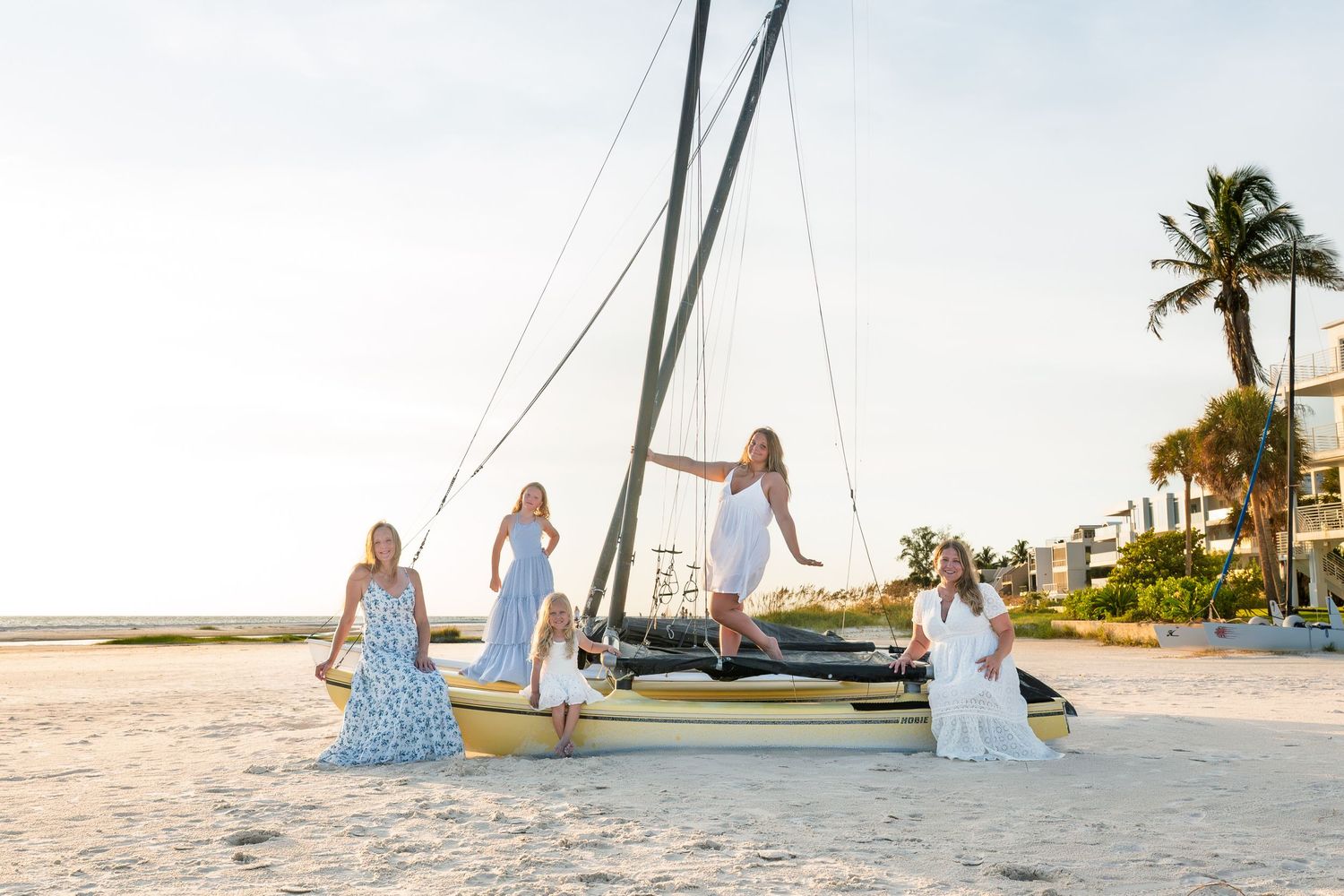 Four women in white and floral dresses pose by a sailboat on a sunny beach with palm trees.