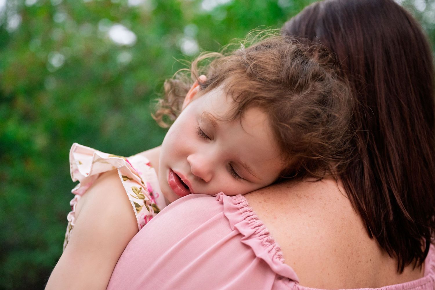 A sleeping baby in a pink outfit rests peacefully outdoors in a natural setting.