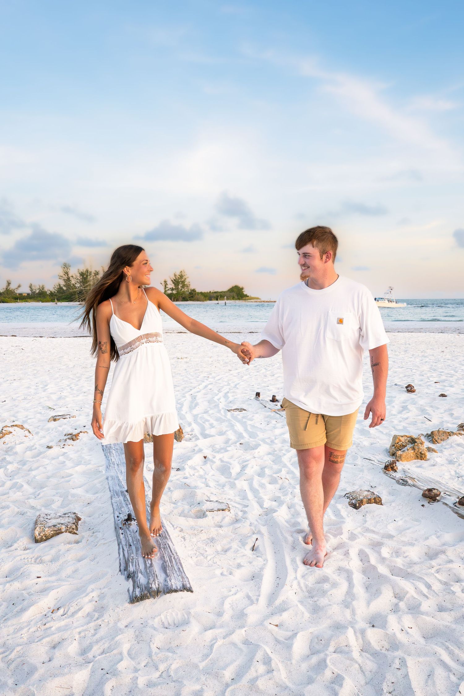 Couple holding hands walking on white sand beach, woman in white dress and man in white shirt and khaki shorts.