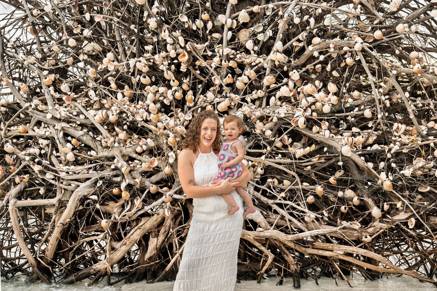 A mother in a white dress holds her baby in front of a decorative twig wall with floral accents outdoors.