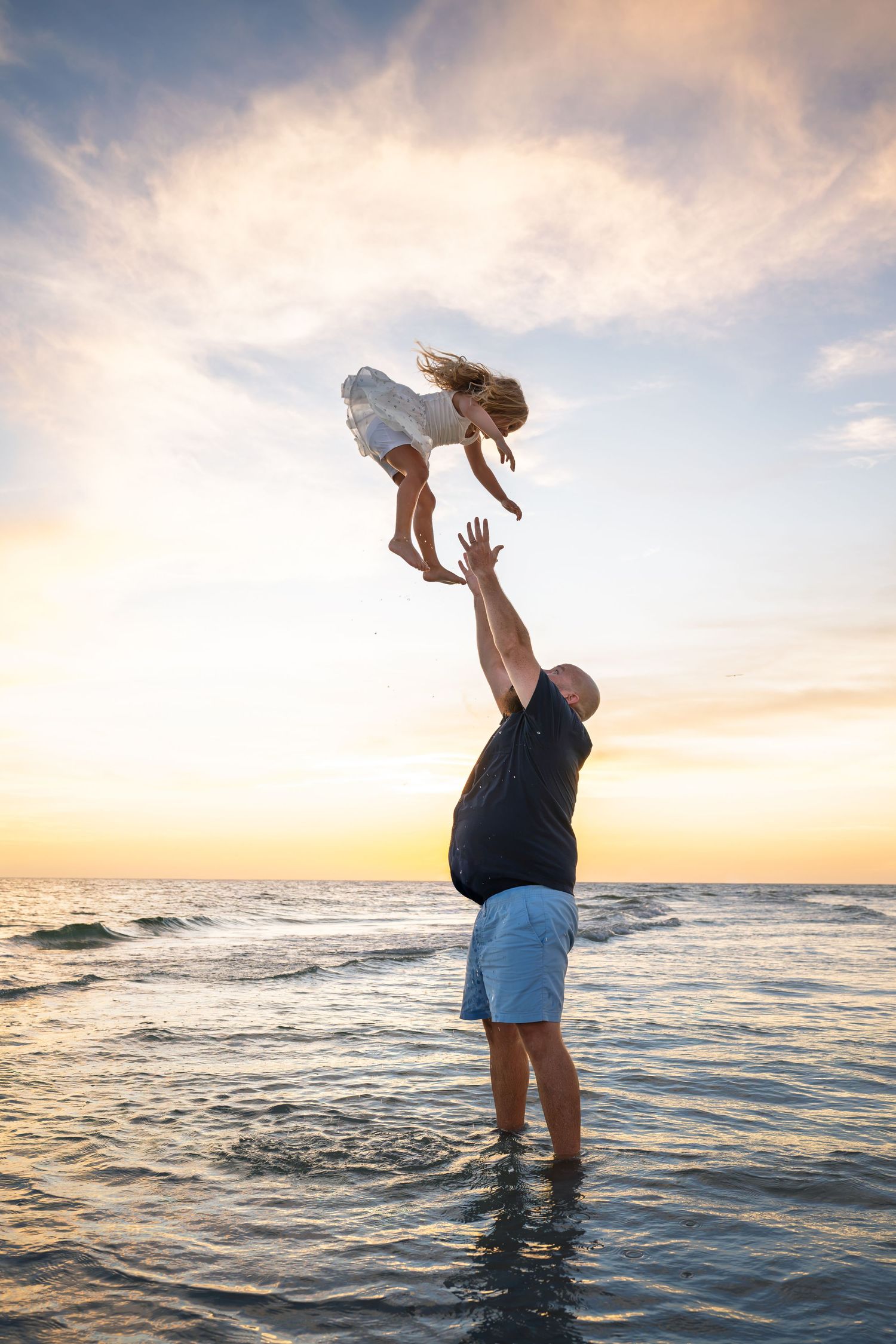 Father joyfully tosses young daughter into the air on a beach at sunset, waves and golden sky in the background.