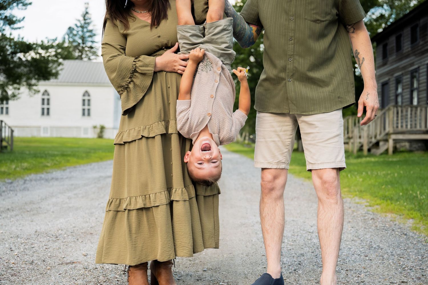 Family photo with parents holding child upside down on gravel path, wearing coordinated green outfits outdoors.