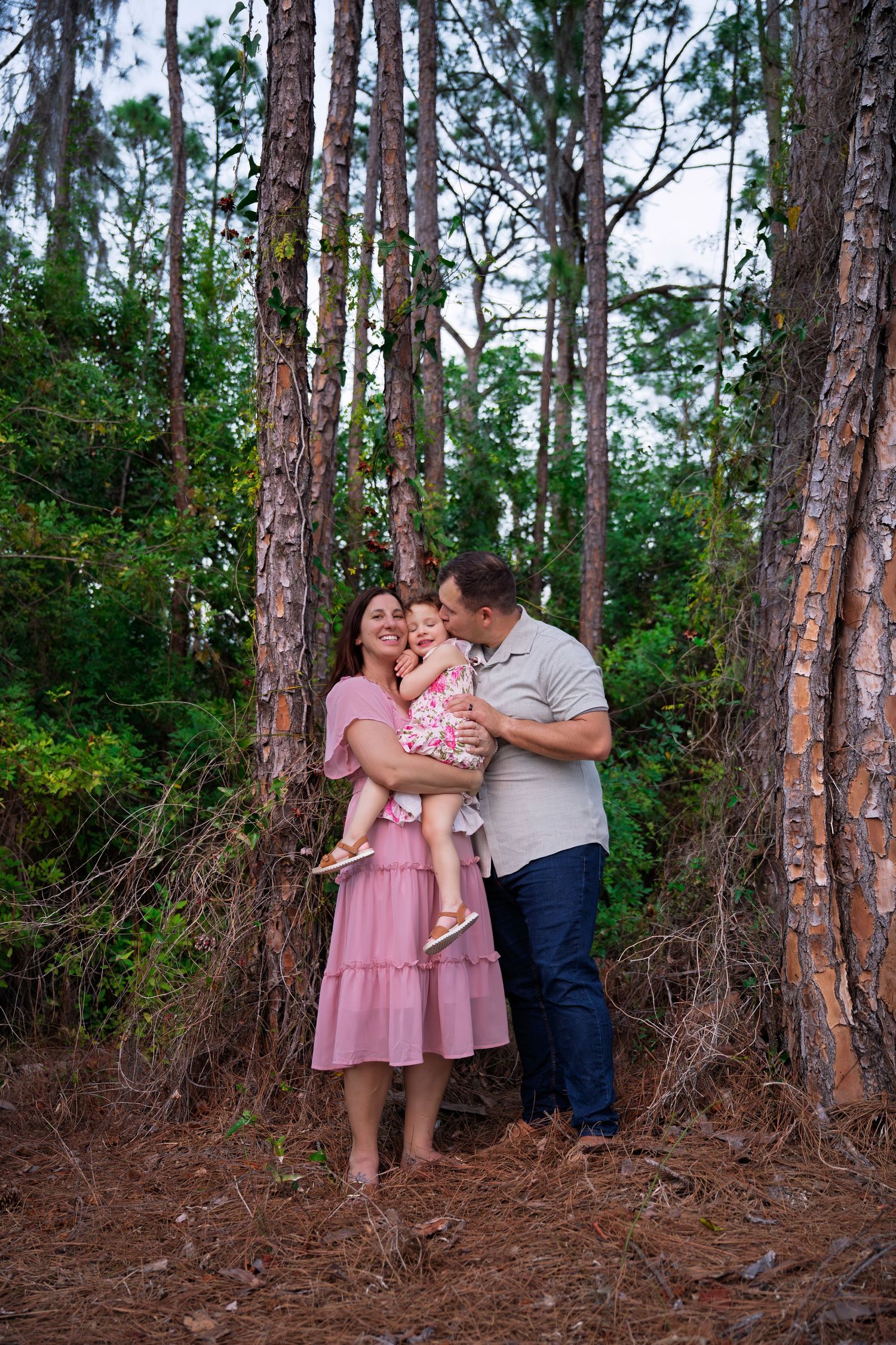 A happy family of three poses in a pine forest, with parents holding their baby girl in a floral outfit.