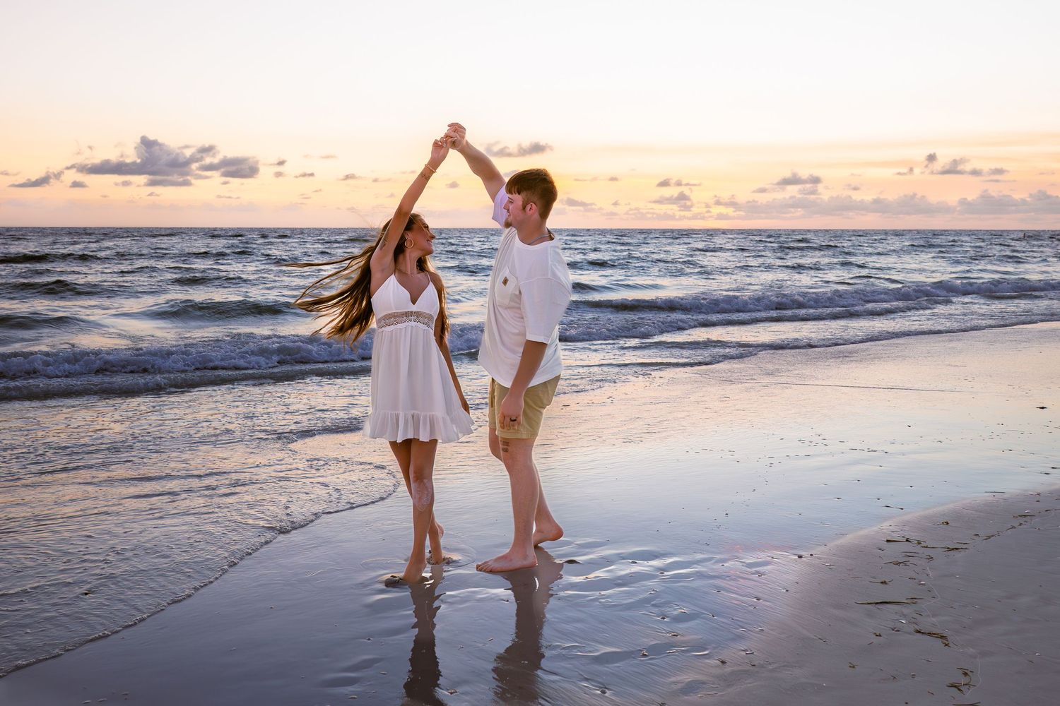 Couple dancing barefoot on a sandy beach at sunset, wearing white outfits, holding hands with ocean waves in background.