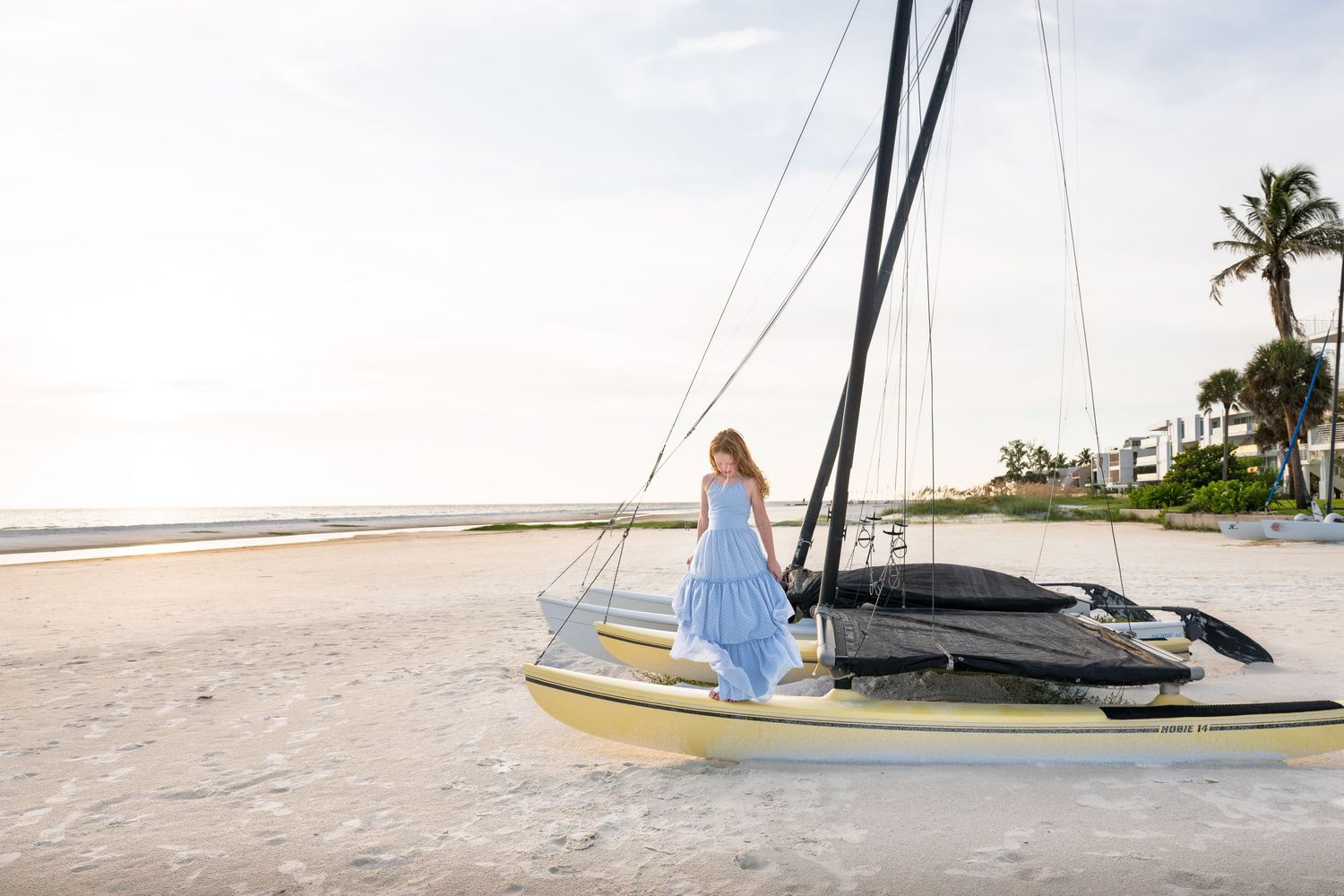 Woman in blue dress posing beside a sailboat on a sandy beach with palm trees in the background.