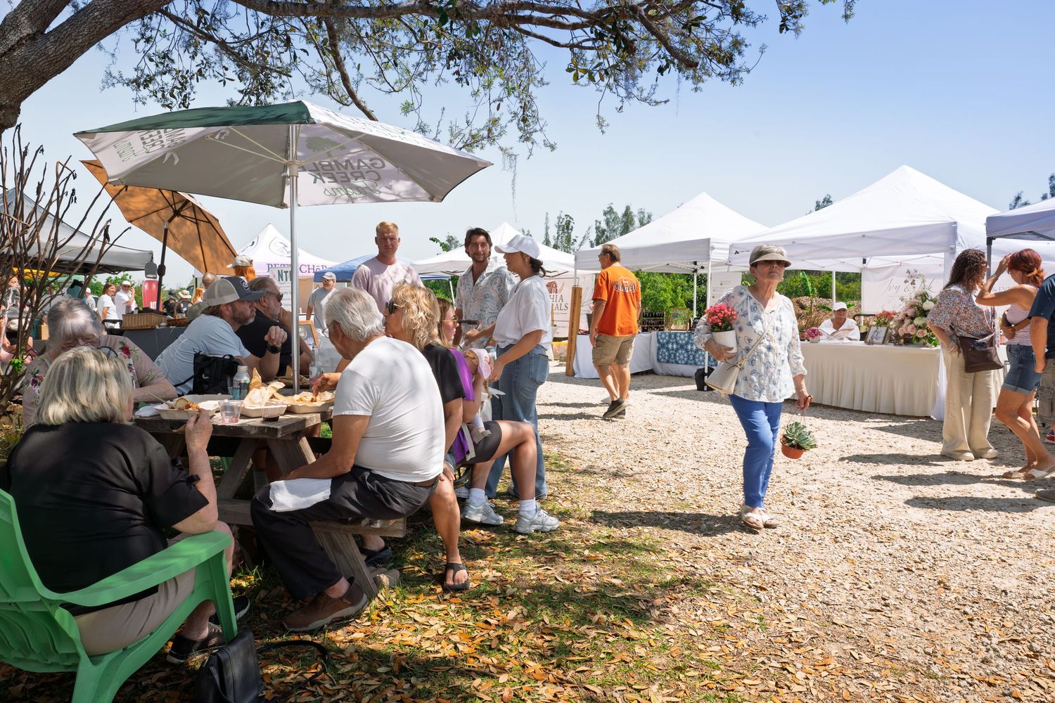 Outdoor farmers market with vendors under white tents, visitors browsing stalls and dining at picnic tables on a sunny day.