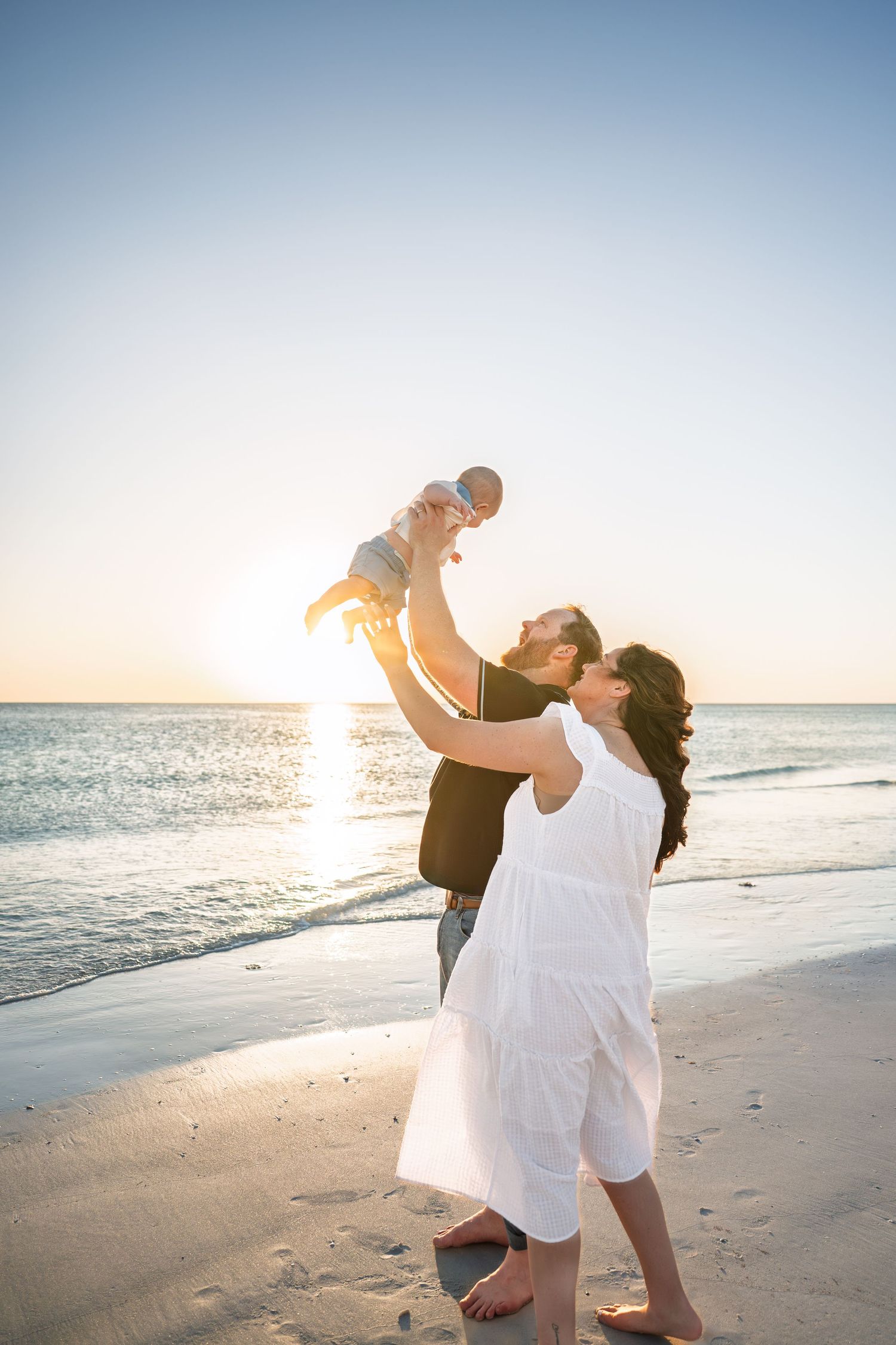 Family enjoying a golden sunset beach moment, father lifting baby in the air while mother watches in white dress.