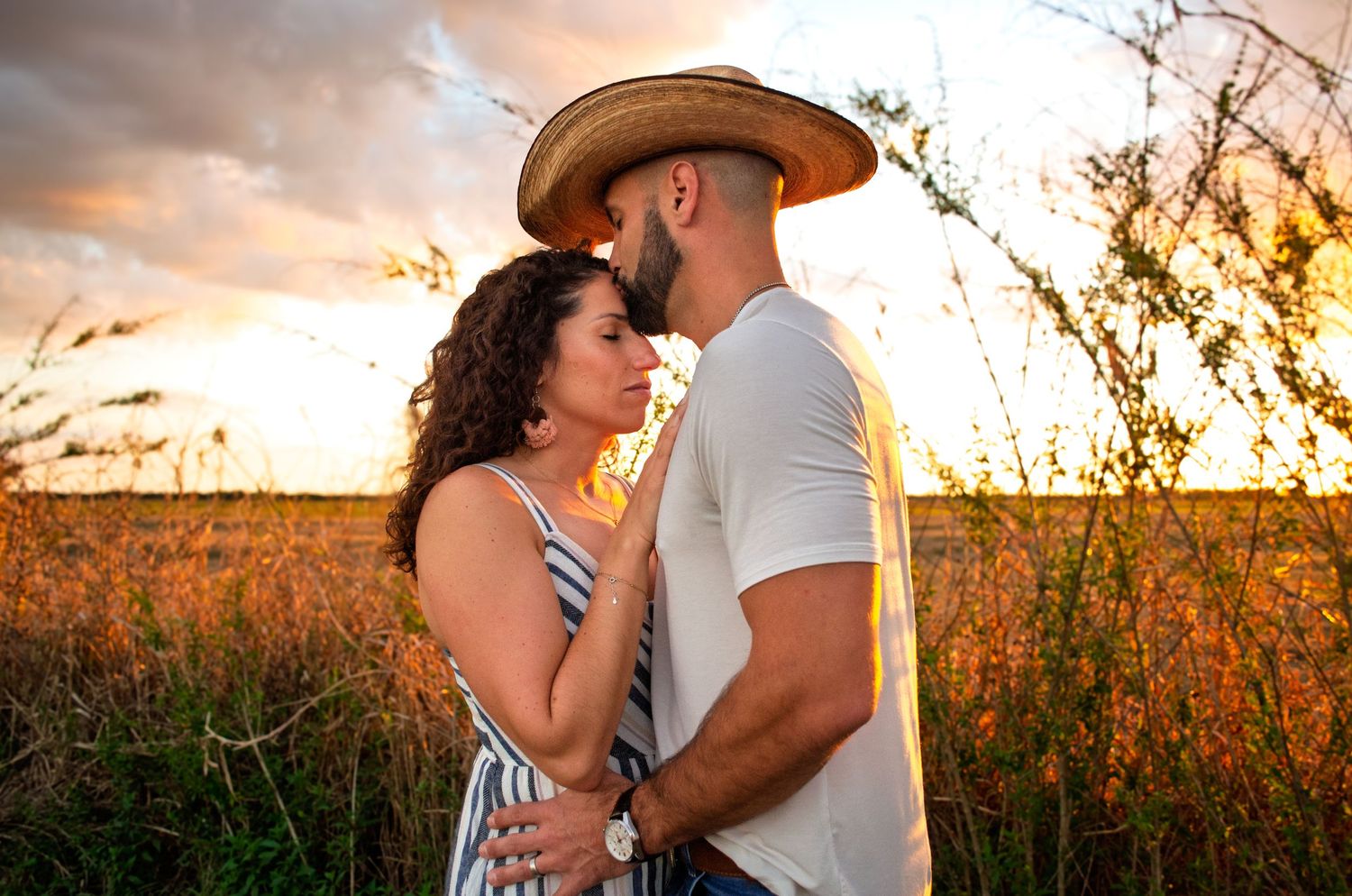 A romantic couple embraces in a golden field at sunset with tall grass and wildflowers surrounding them.