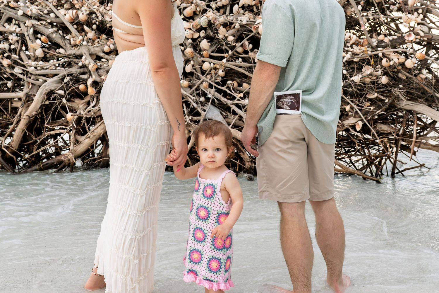 Family of three stands on beach shore, parents holding toddler girl's hands, mother showing ultrasound photo.