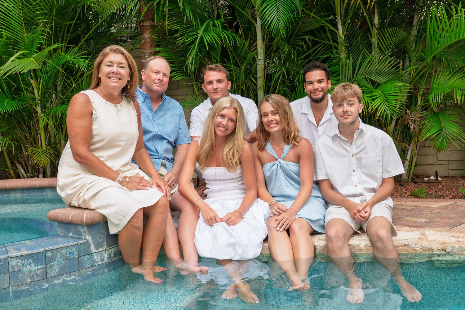 Family of seven smiling together poolside surrounded by tropical greenery on a sunny day.