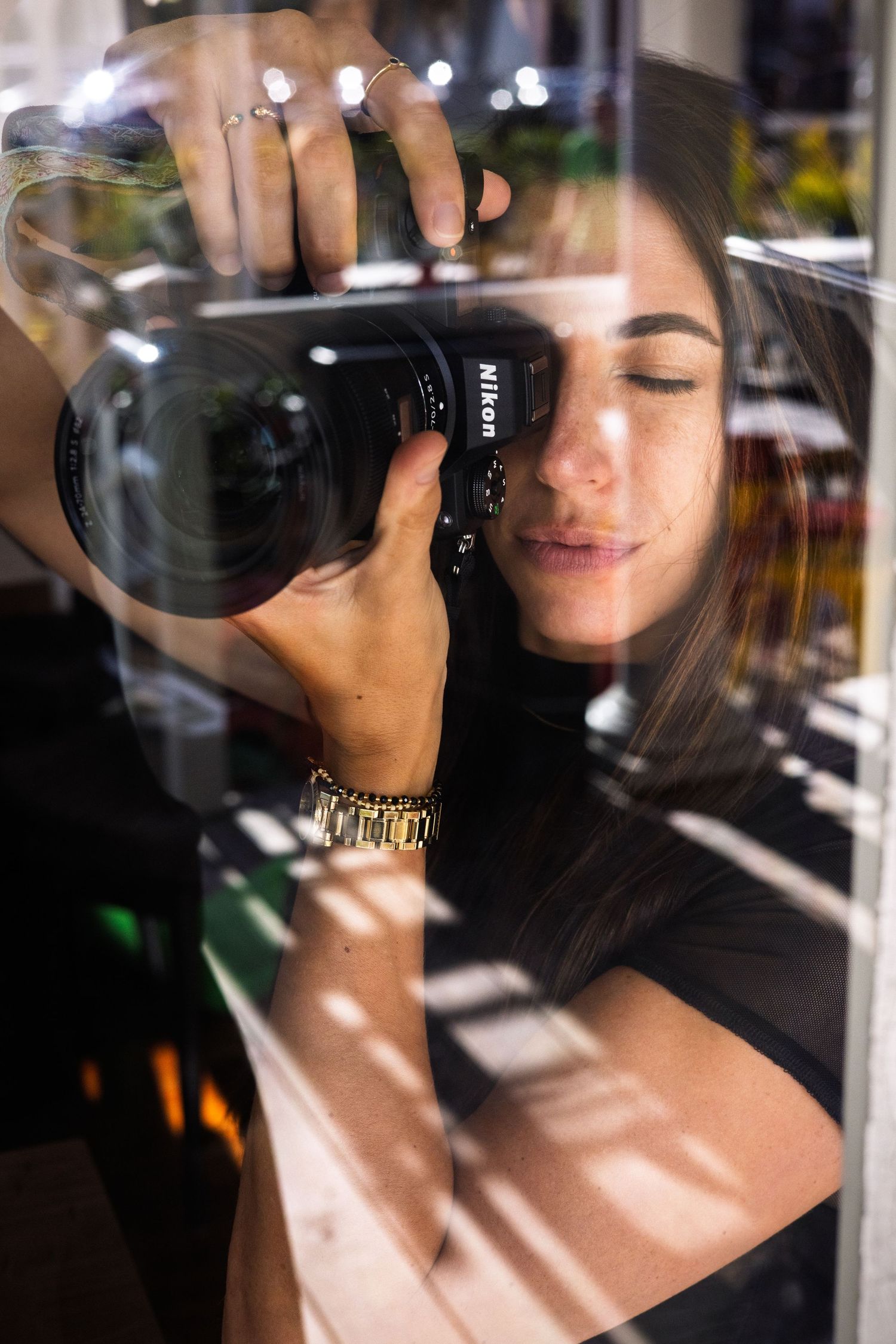 Young woman photographing with a Nikon DSLR camera, reflected in glass, wearing a gold bracelet and rings.