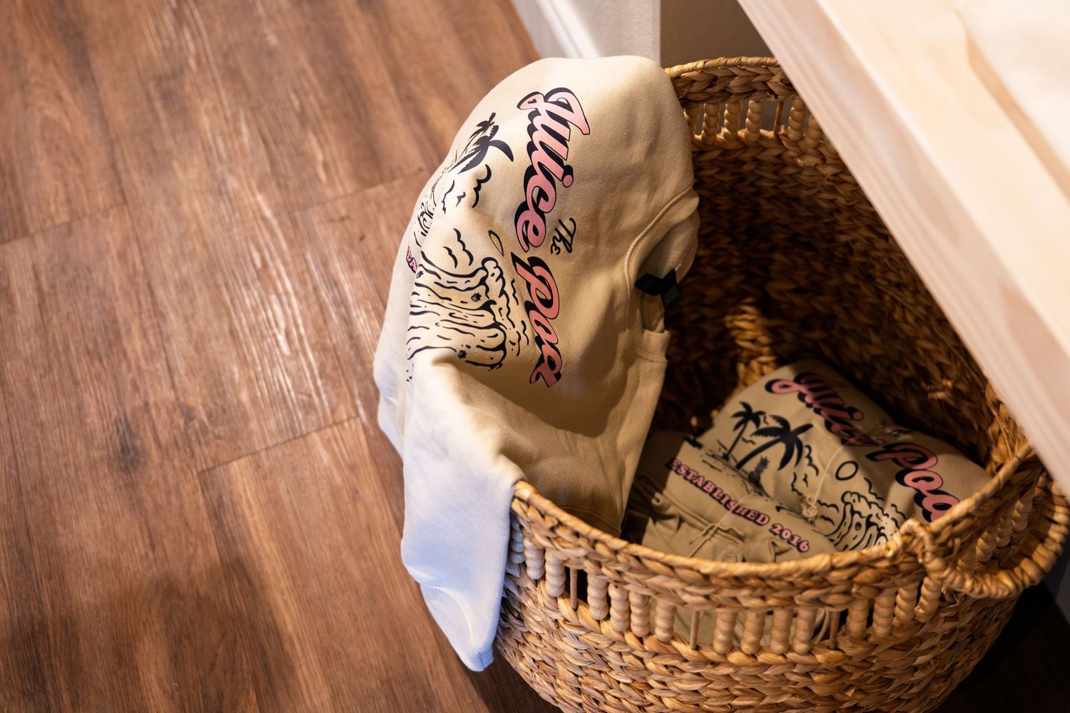 Wicker basket with folded clothes and decorative pillow on hardwood floor.