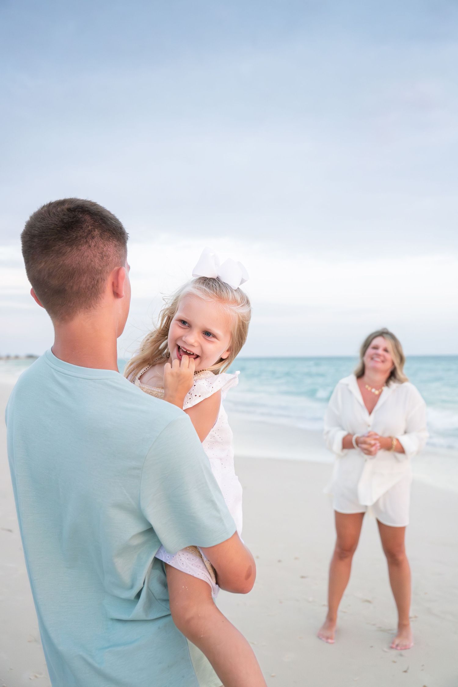 Family enjoying a beach day, father holding laughing young girl in white dress while mother smiles in background.