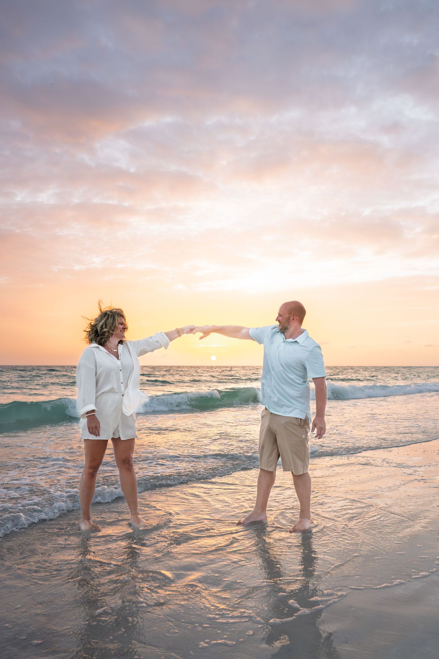 Couple holding hands at sunset on a beach, waves crashing behind them, wearing white and khaki outfits.