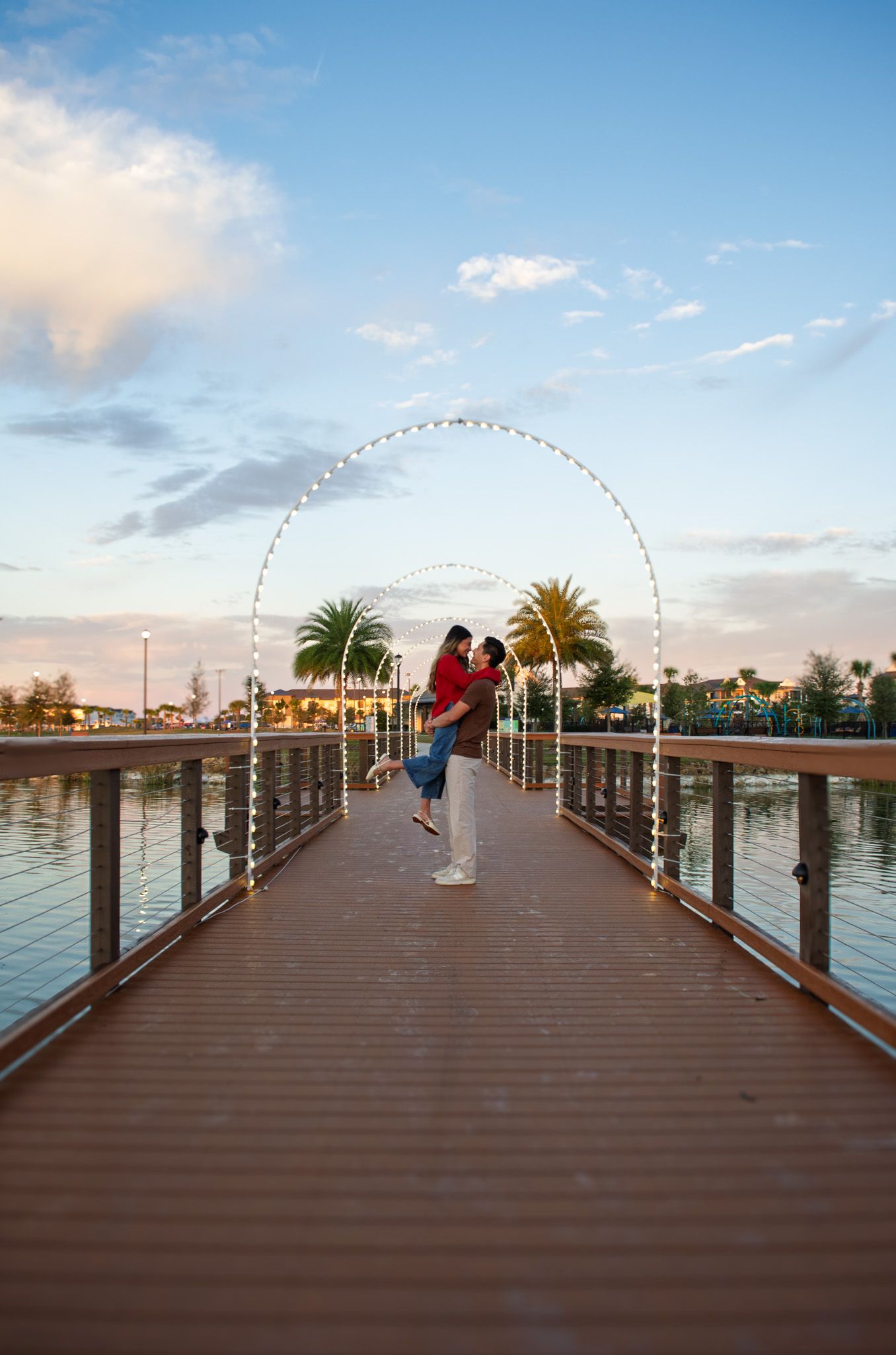 A wooden boardwalk pier extends over calm waters at sunset with palm trees and buildings in the background.