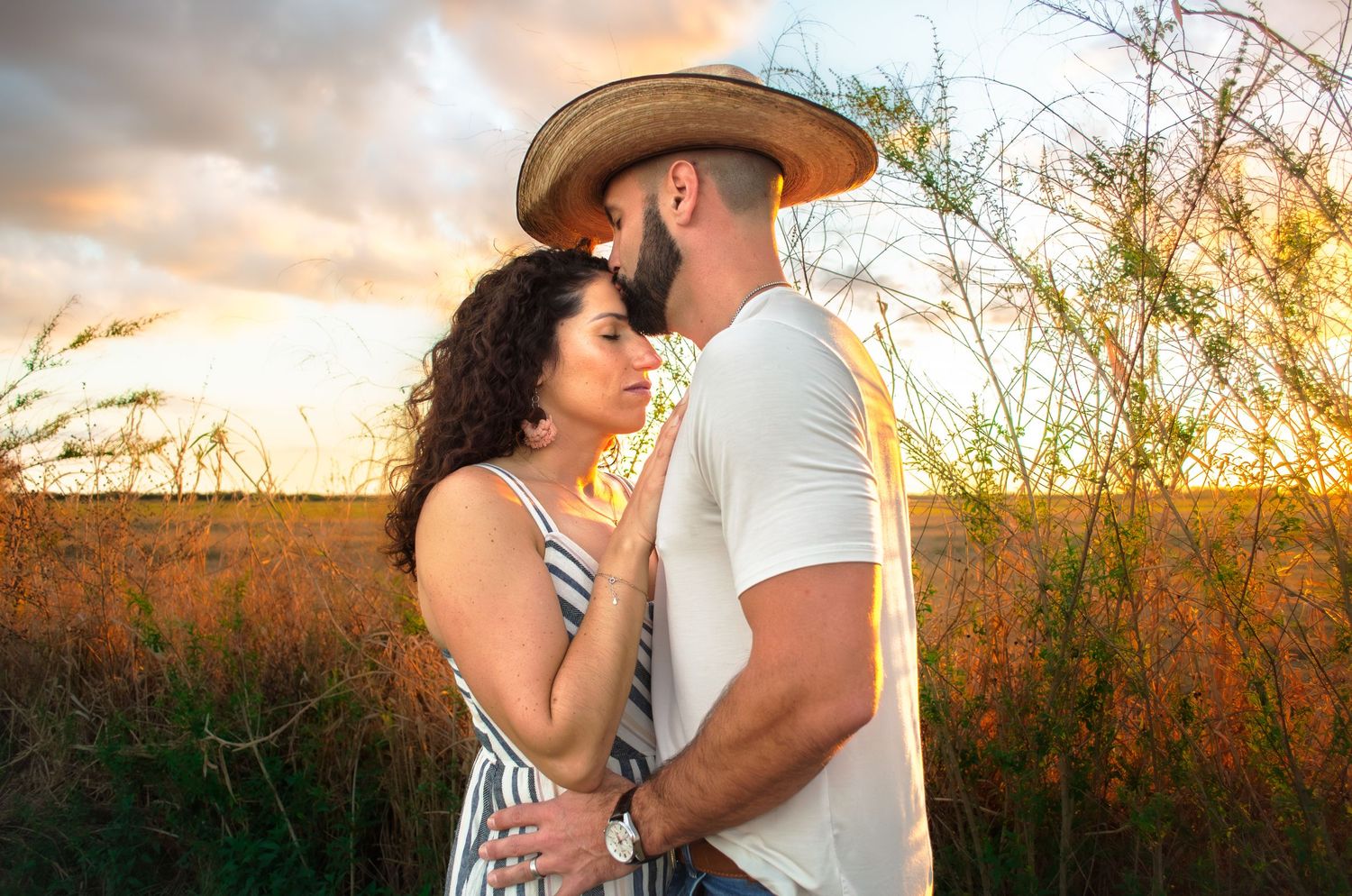 A couple shares a romantic moment at sunset in a golden field, the man wearing a straw hat embracing a curly-haired woman.