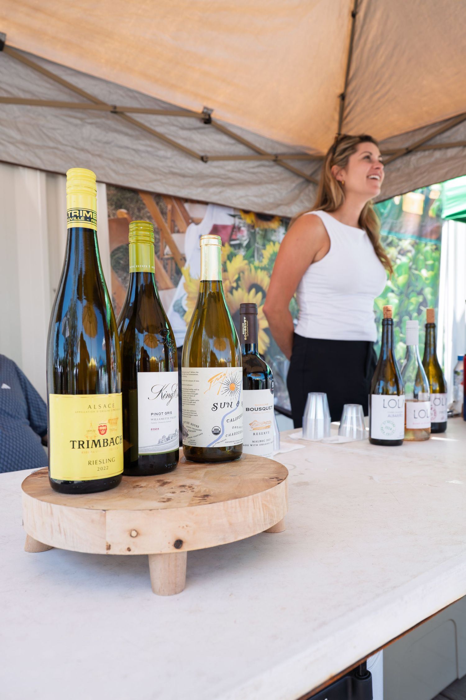 Wine tasting event with Trimbach Riesling bottles on wooden stand, woman smiling in background under tent.