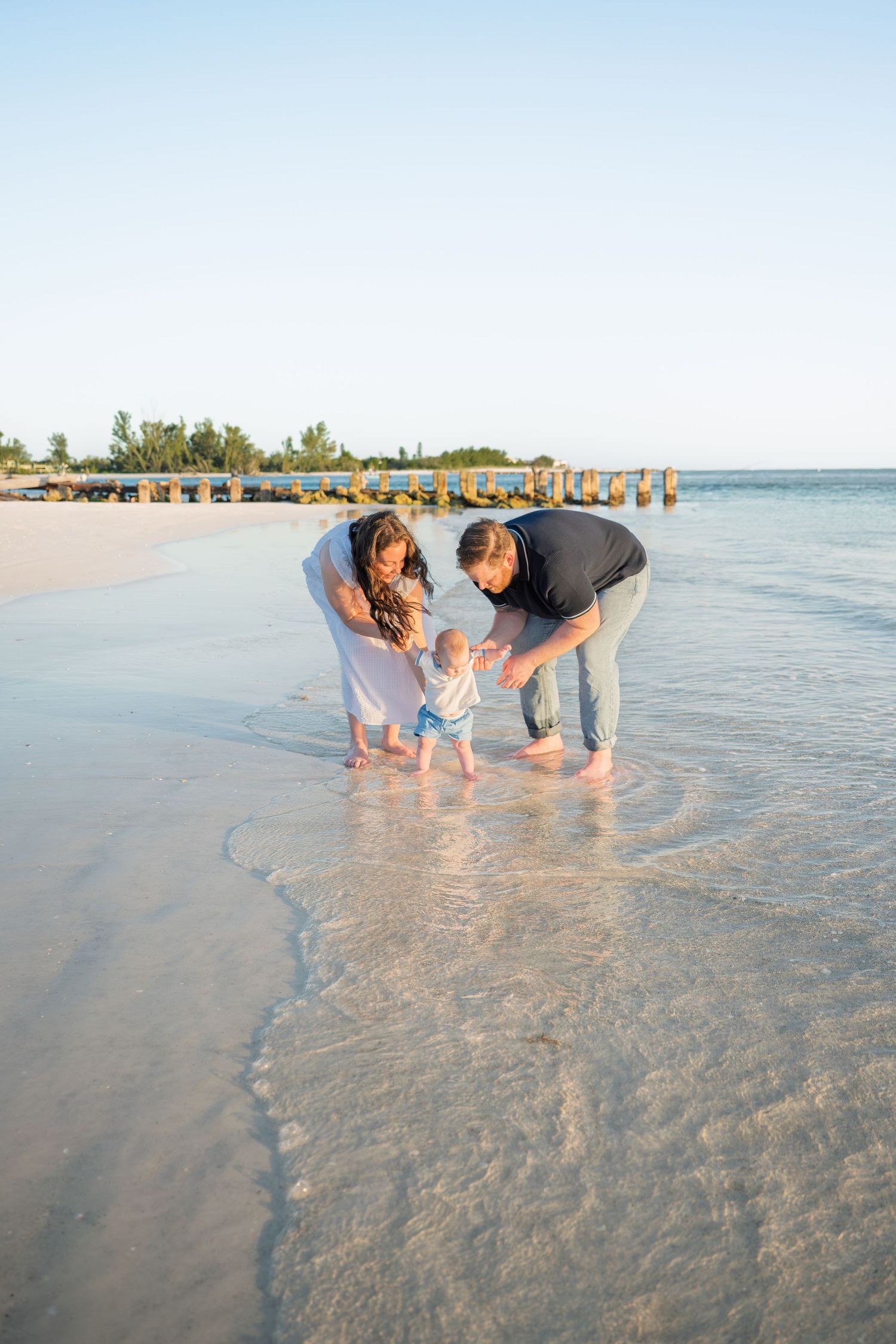 Family with baby enjoying shallow ocean waves on a sandy beach near a wooden pier at sunset.