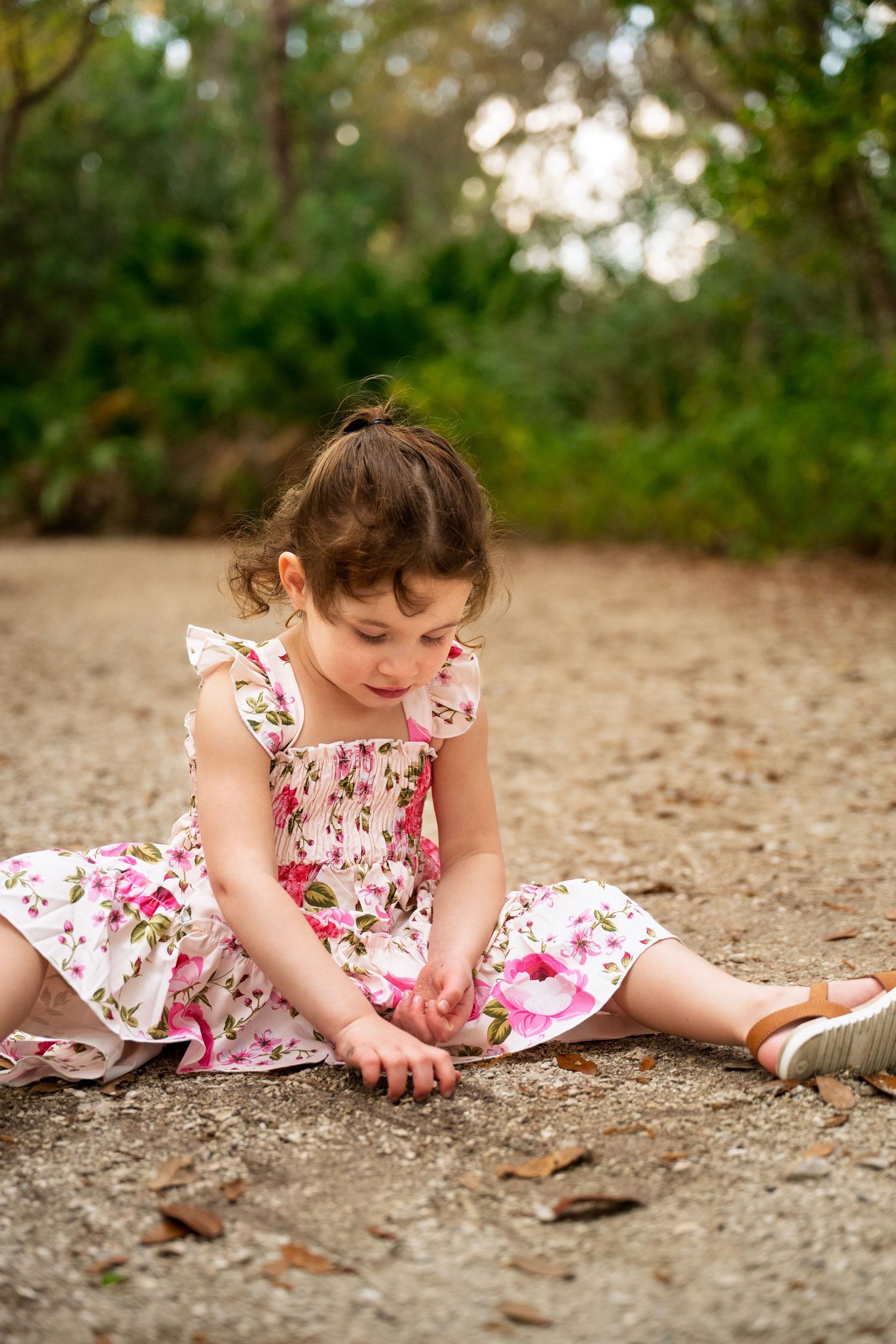 A child in a floral dress sits on sandy ground examining something with curiosity in a natural outdoor setting.
