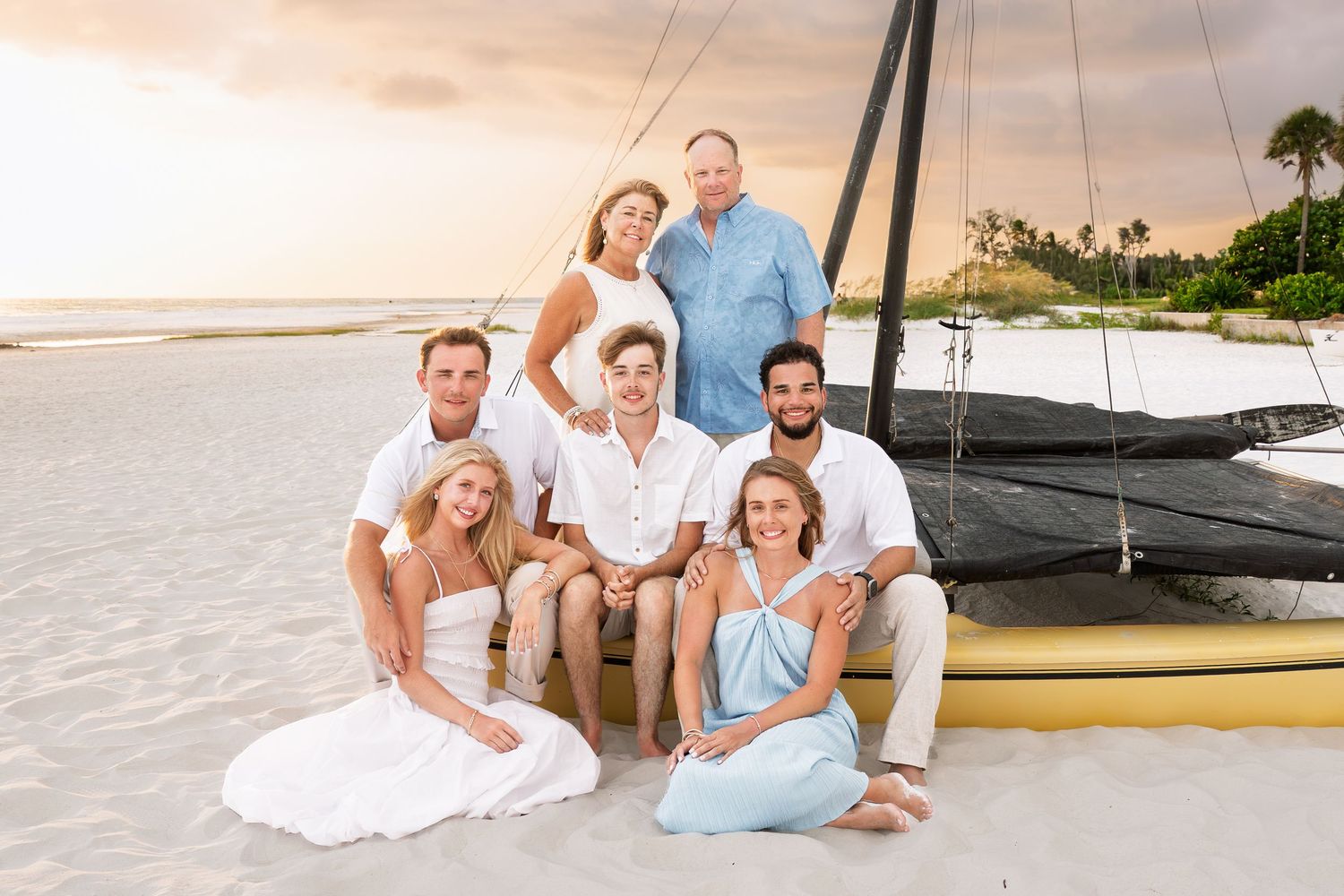 Family of seven posing together on a tropical beach near a sailboat at sunset, dressed in white and light blue outfits.