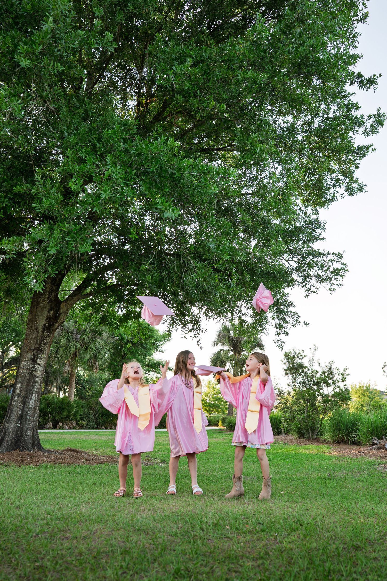 Three girls in pink graduation gowns tossing caps in the air outdoors.