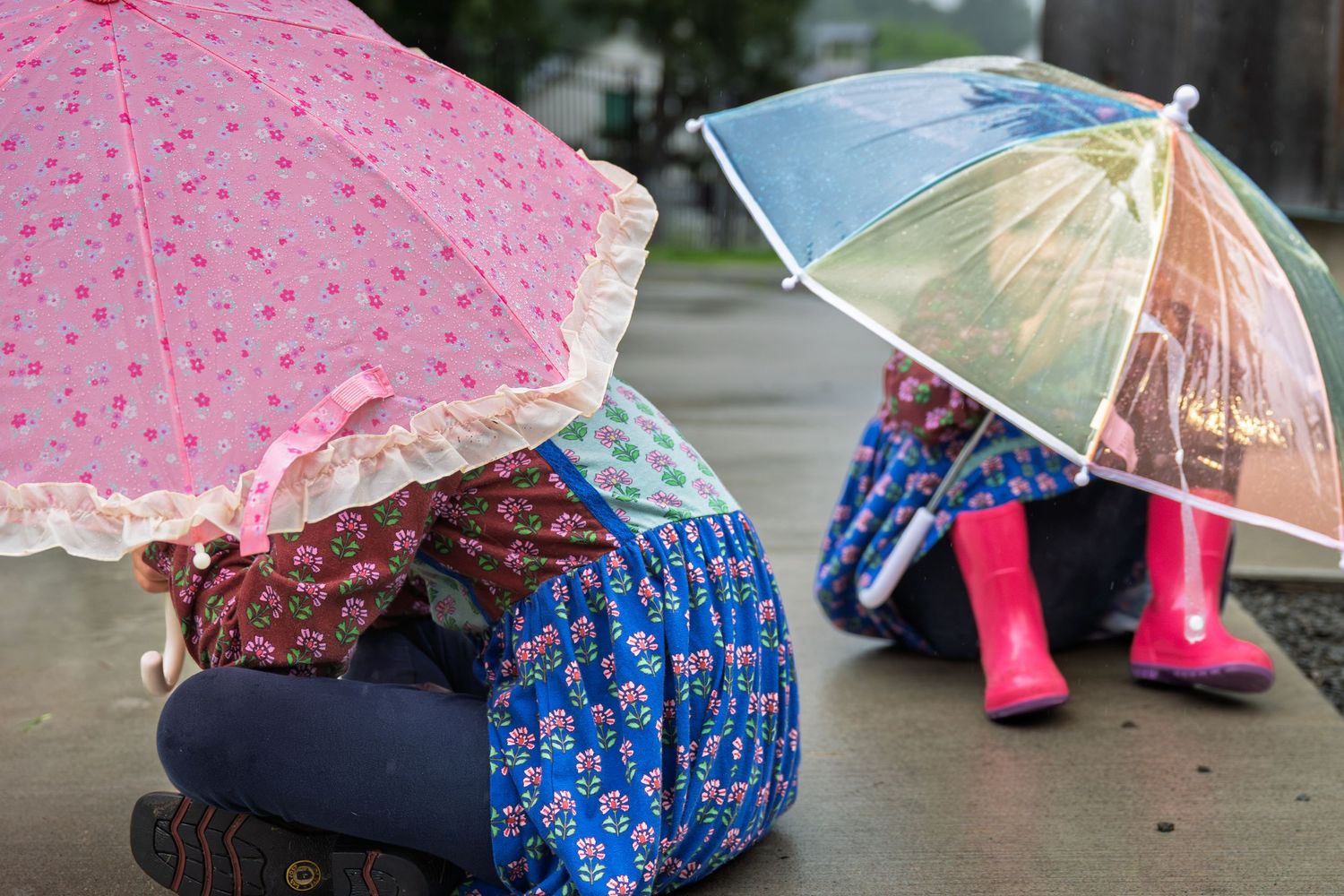 Two children sitting on wet pavement, sheltering under colorful umbrellas in the rain, wearing rain boots and floral outfits.