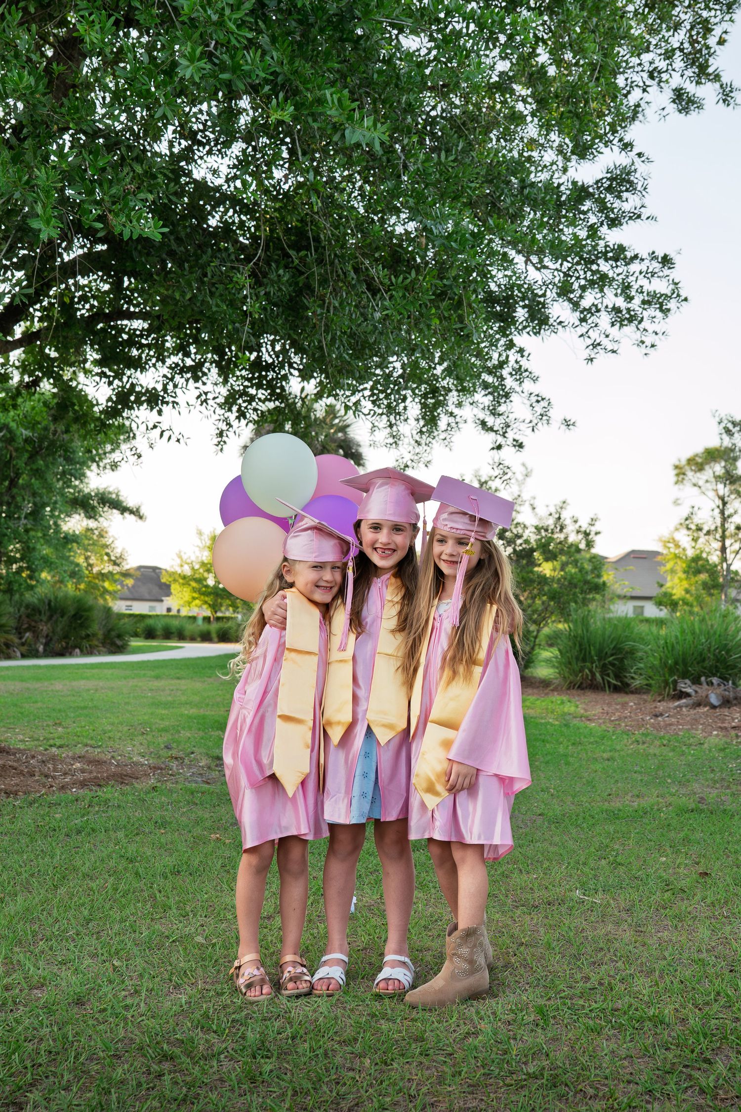 Three girls in pink graduation caps and gowns holding balloons outdoors.