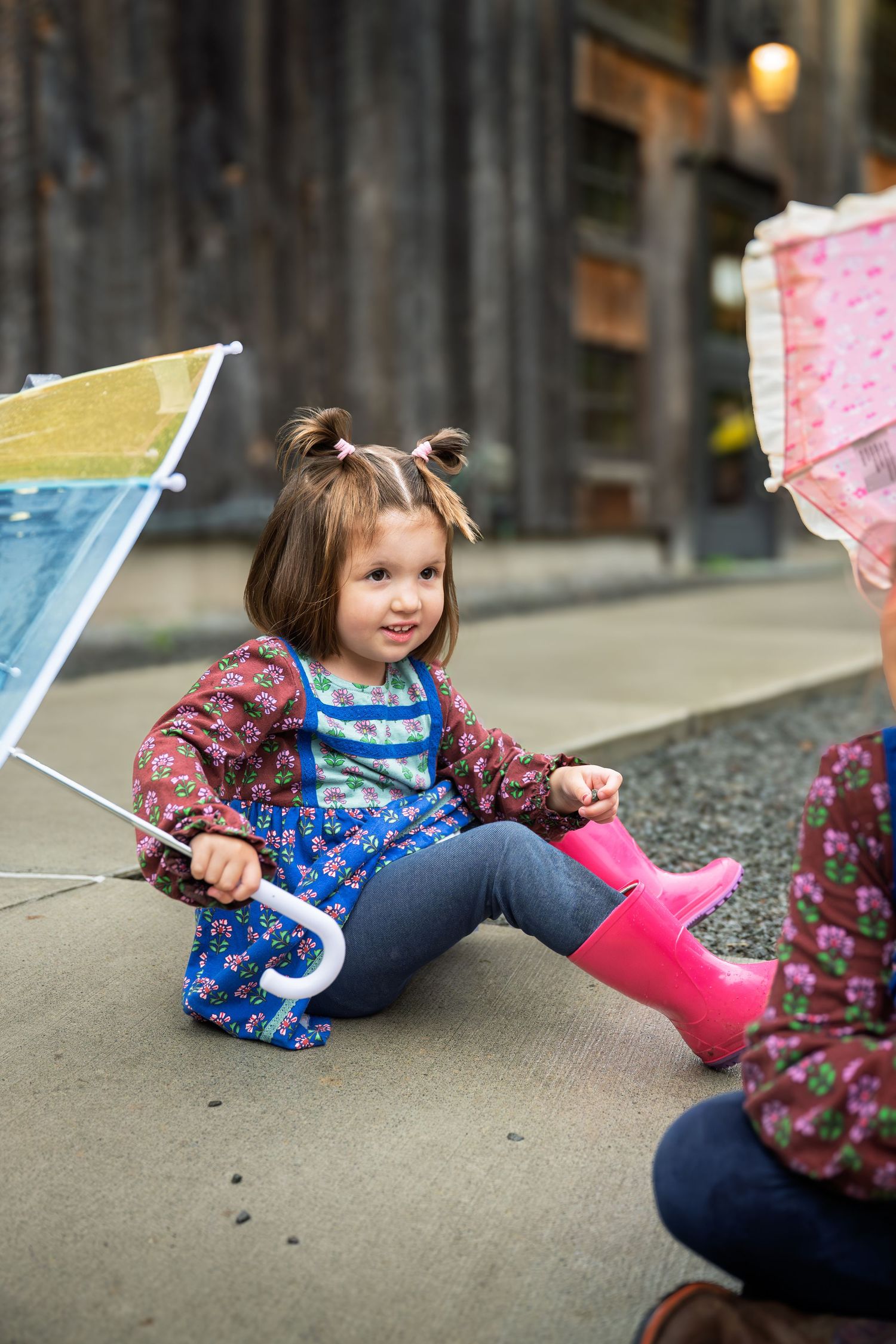 Toddler girl in pink rain boots and floral dress sits on pavement holding a colorful umbrella.