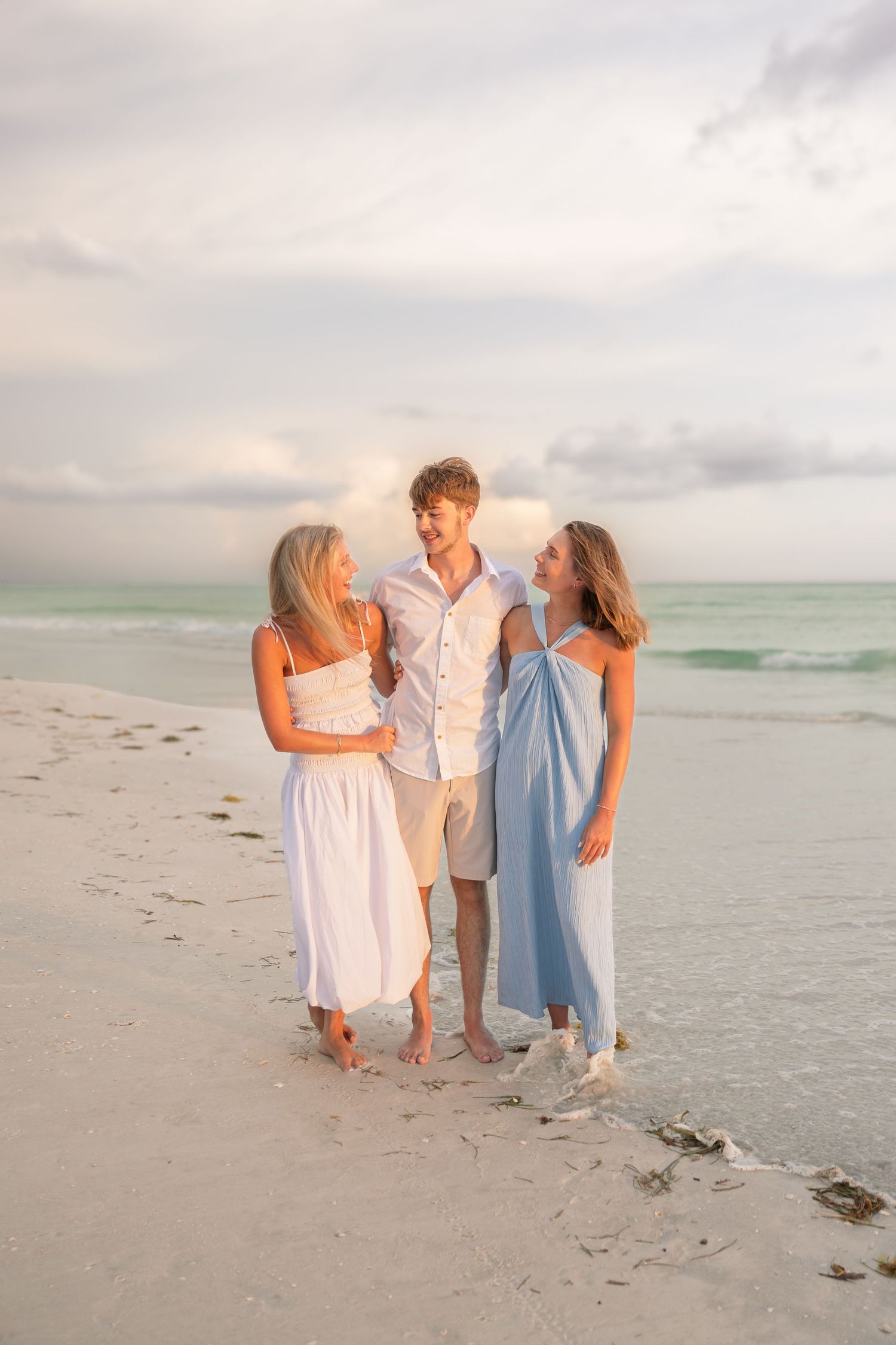 Three people in white and blue outfits walking barefoot on a sandy beach at sunset.