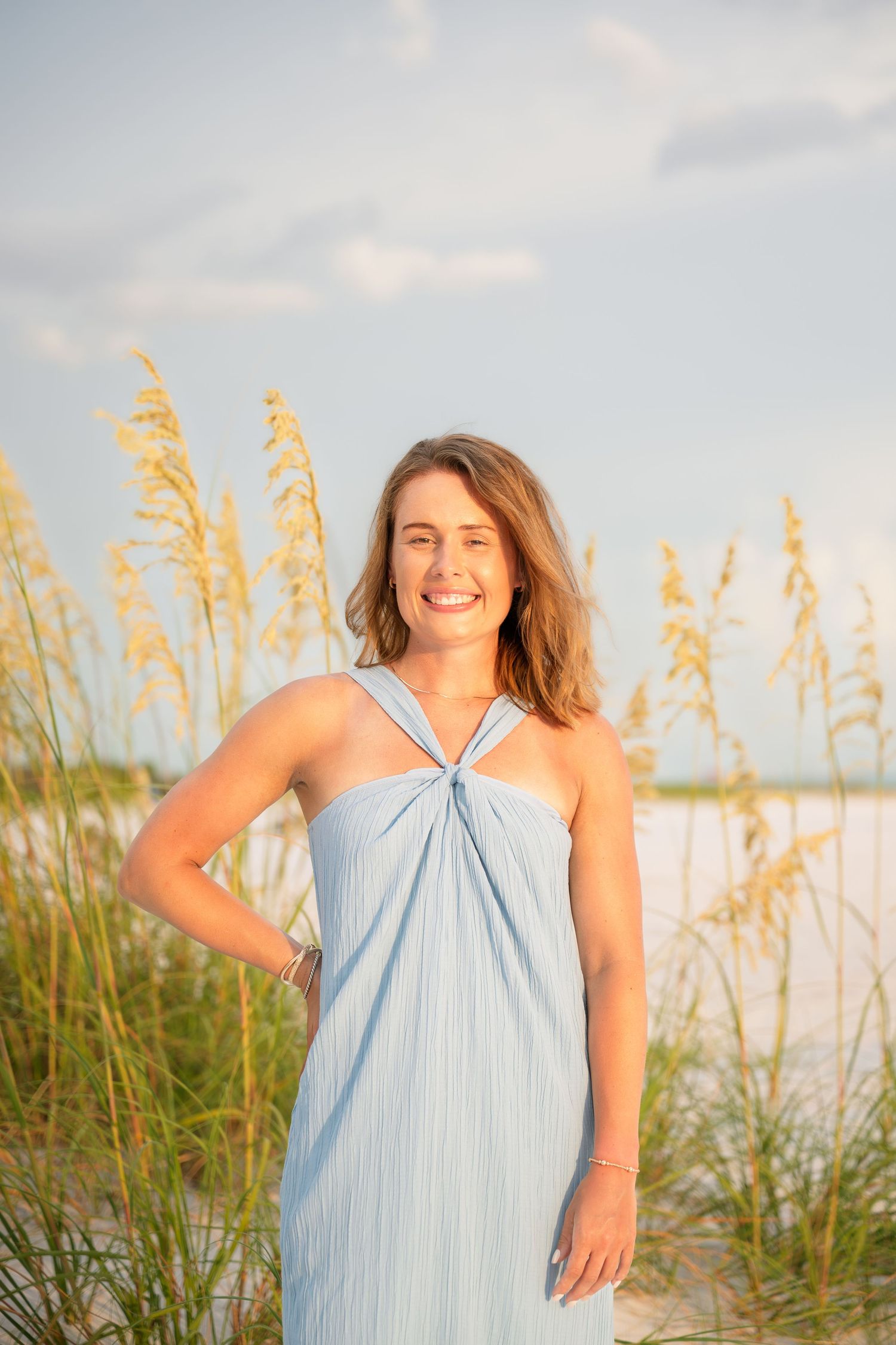 Smiling woman in light blue sundress stands among sea oats on a sunny beach with blue sky.