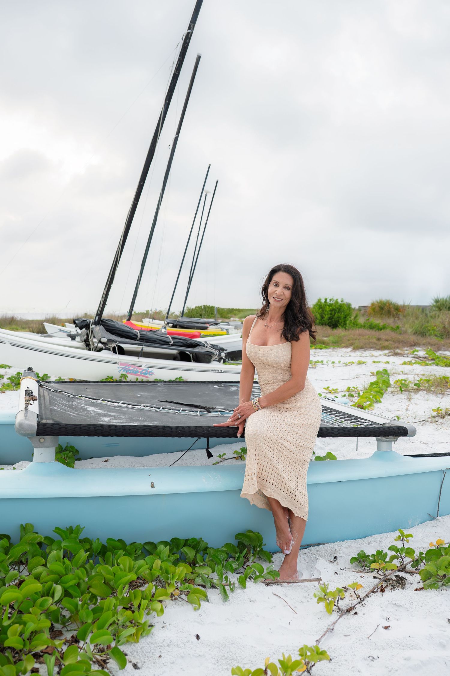 Woman in cream dress poses beside blue catamaran on sandy beach with sailboats and green foliage in background.