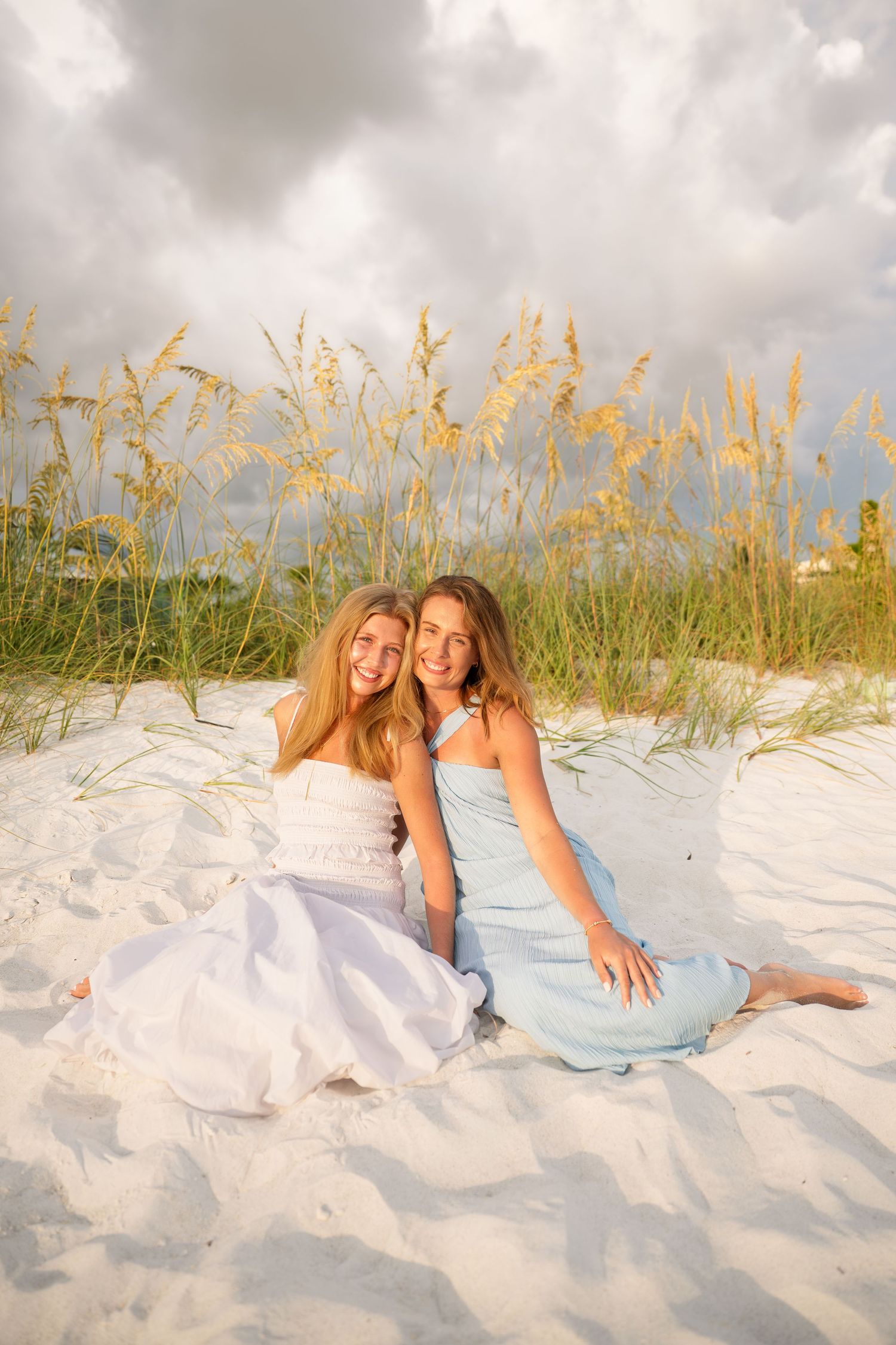 Two smiling girls in pastel dresses sit together on white sand beach with sea oats and cloudy sky.